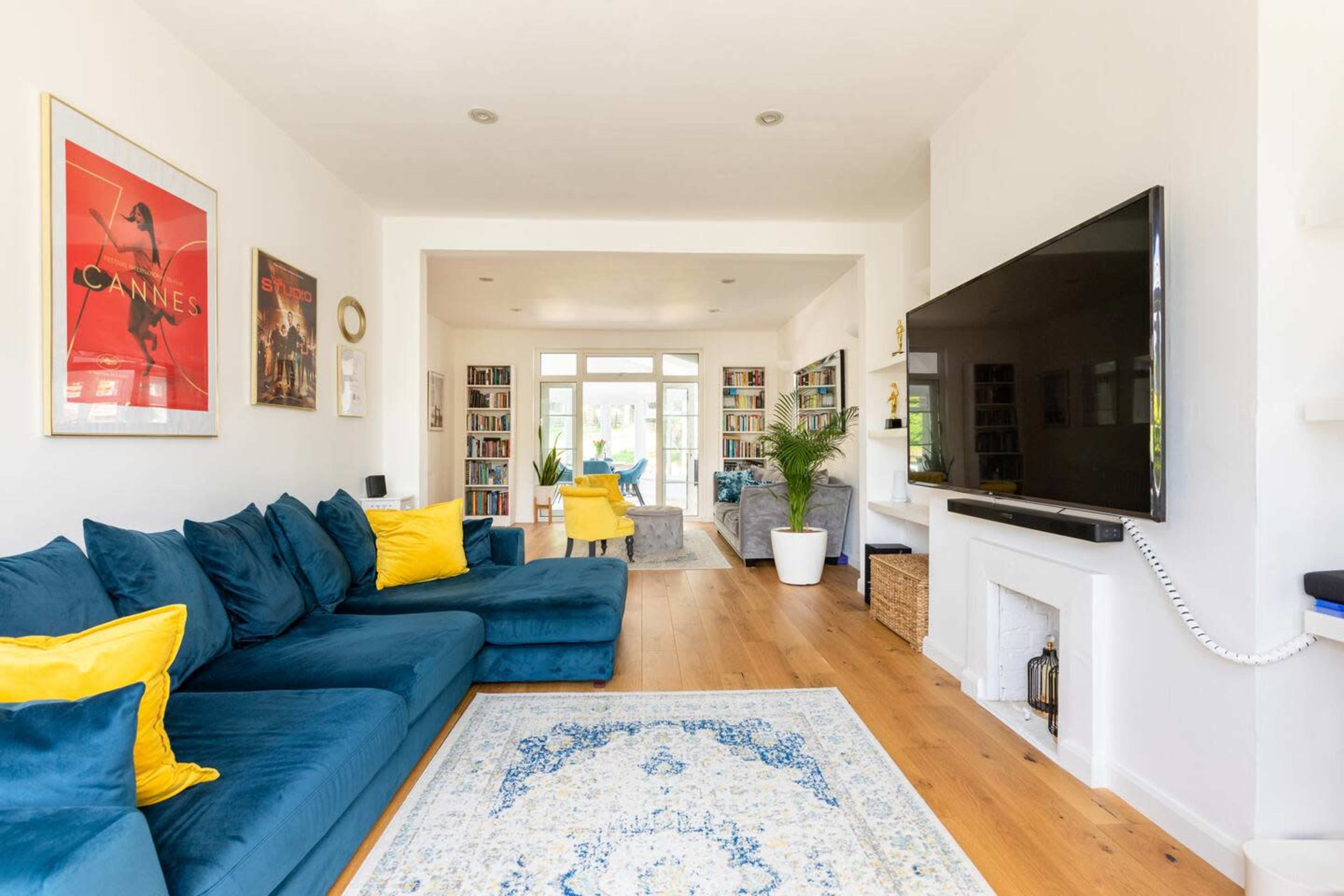 The living room features a blue sectional sofa with yellow cushions, a large television mounted on the wall, and shelves filled with books.