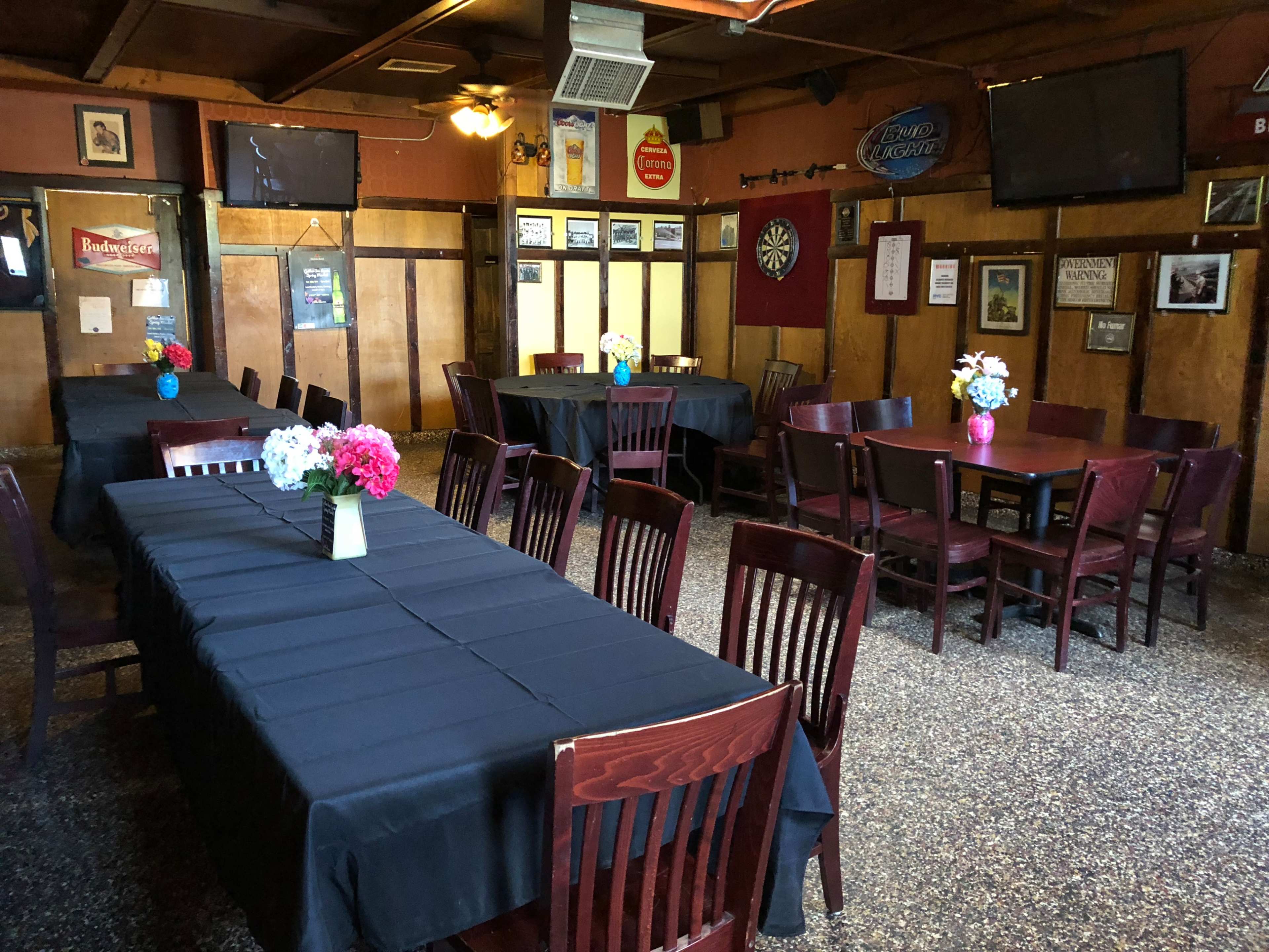 The interior of a restaurant features several tables with dark cloths and centerpieces of flowers, arranged in a spacious, dimly lit area.