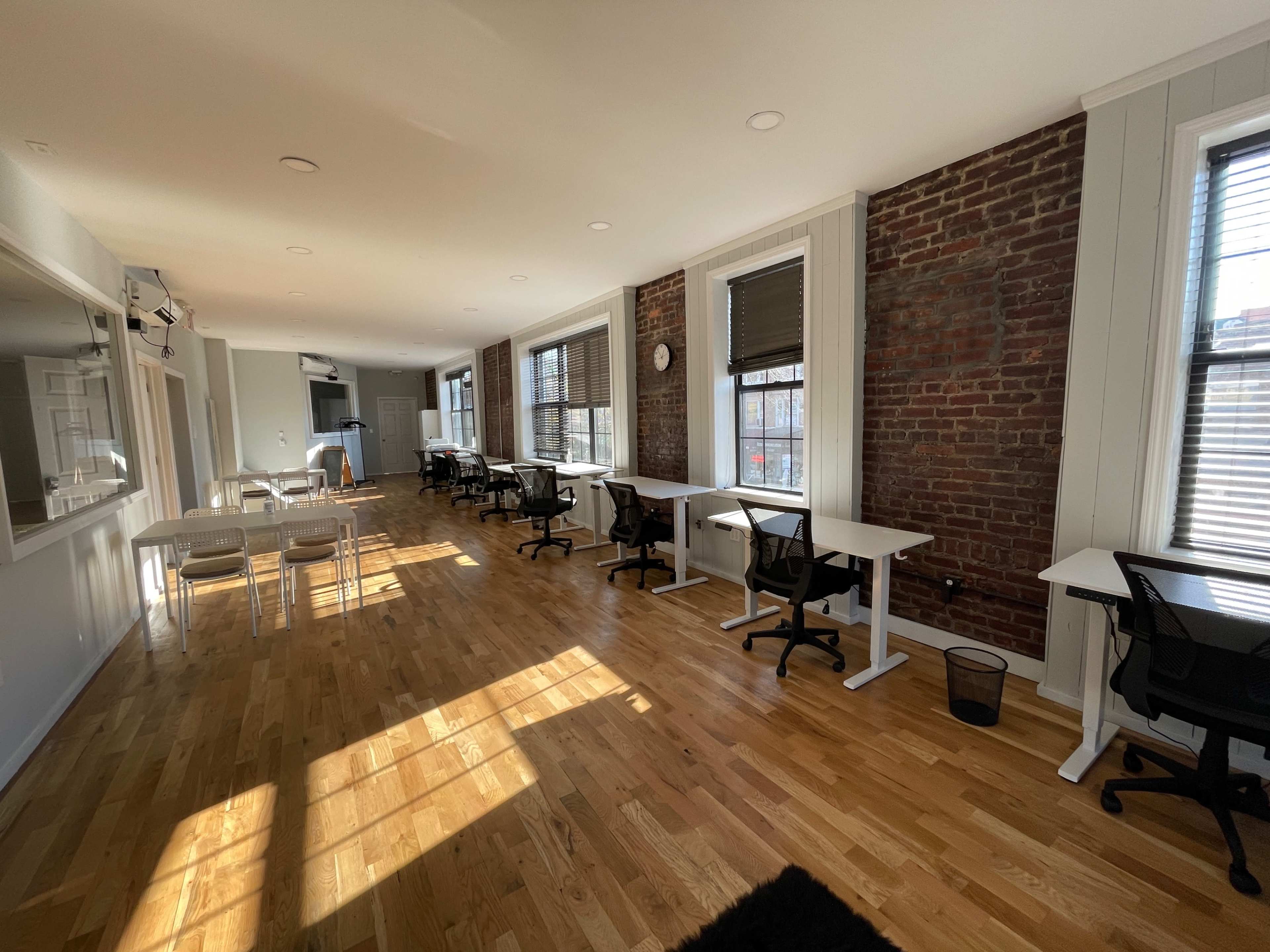 A spacious office interior with wooden floors, white desks, and black office chairs arranged along large windows, illuminated by natural light.