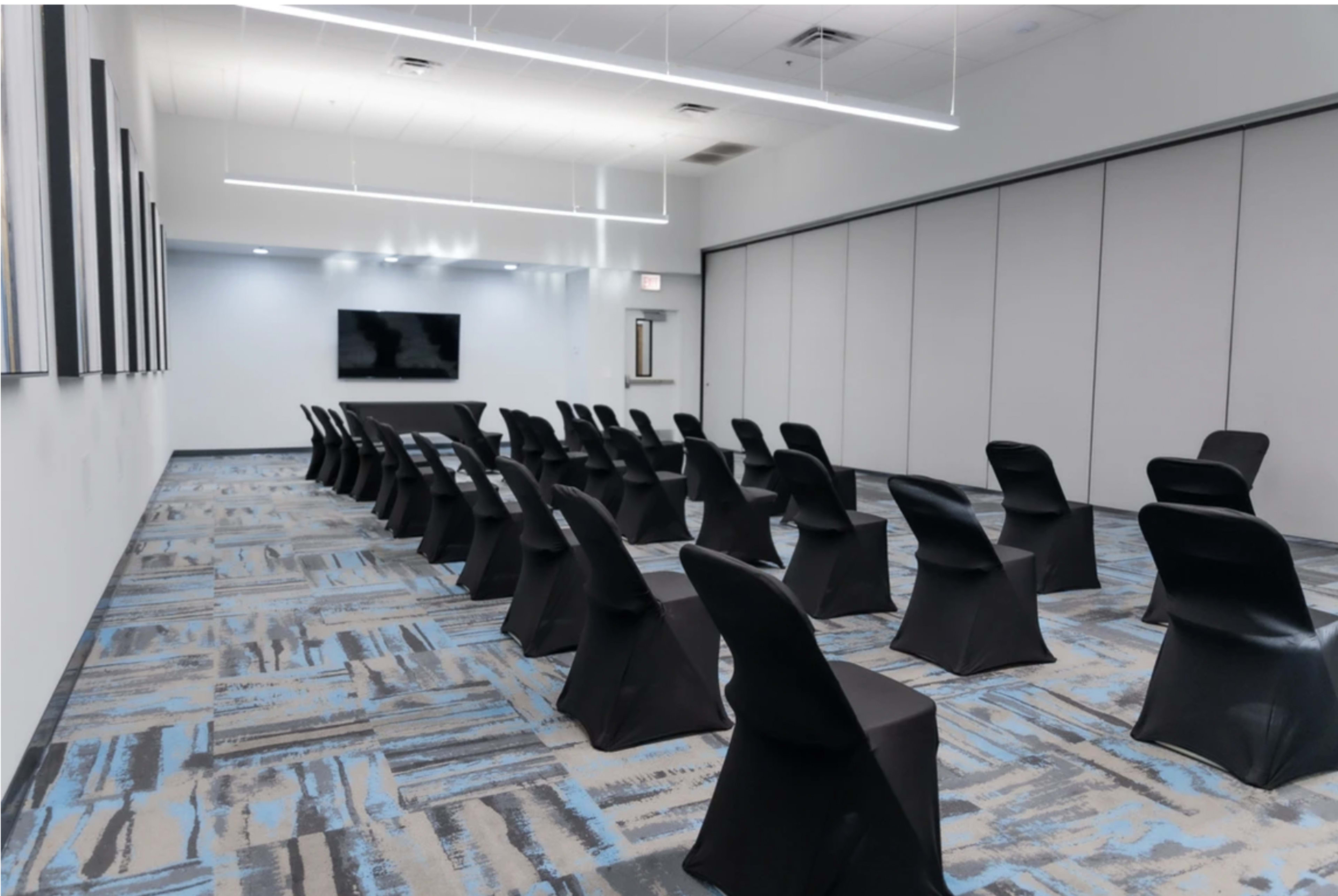 A conference room with rows of black chairs arranged facing a wall-mounted screen, set against a gray and blue carpet.