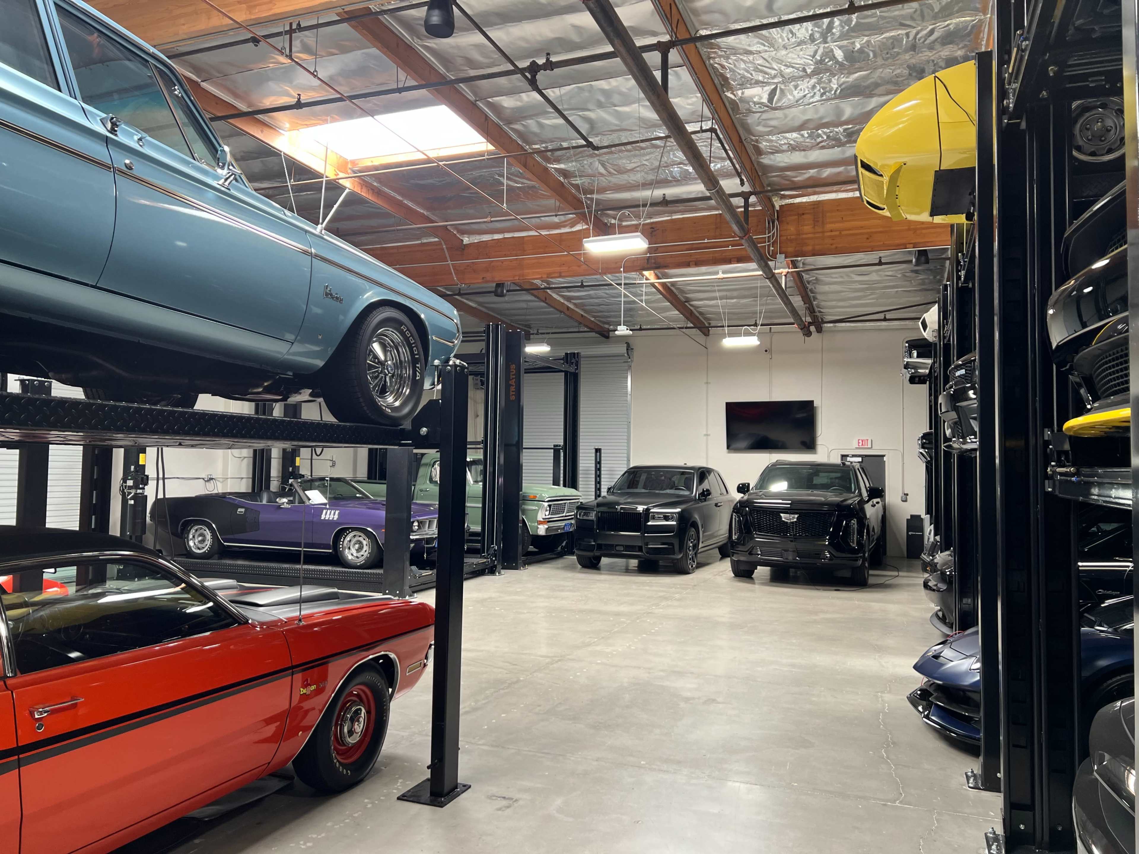 The image shows a spacious garage with multiple classic and modern cars, including a blue car on a lift, parked alongside others in organized rows.