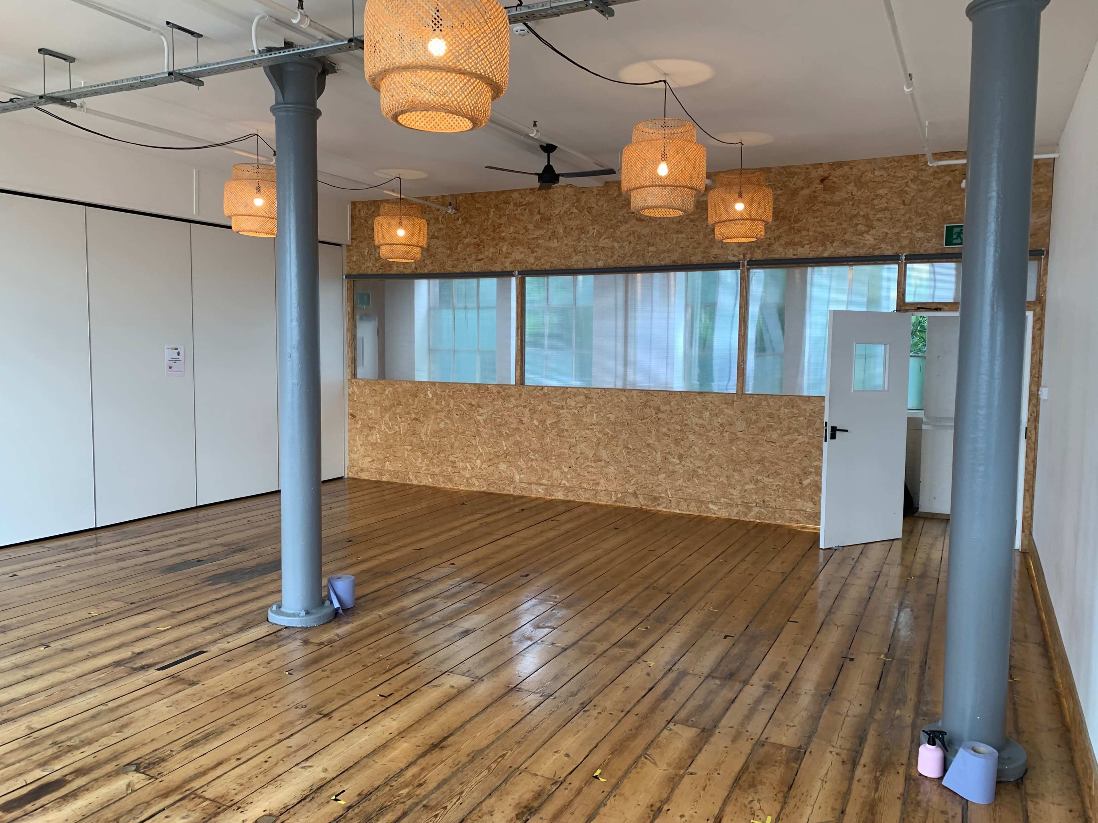 A spacious room with wooden floors, exposed beams, large windows, and pendant light fixtures hanging from the ceiling.