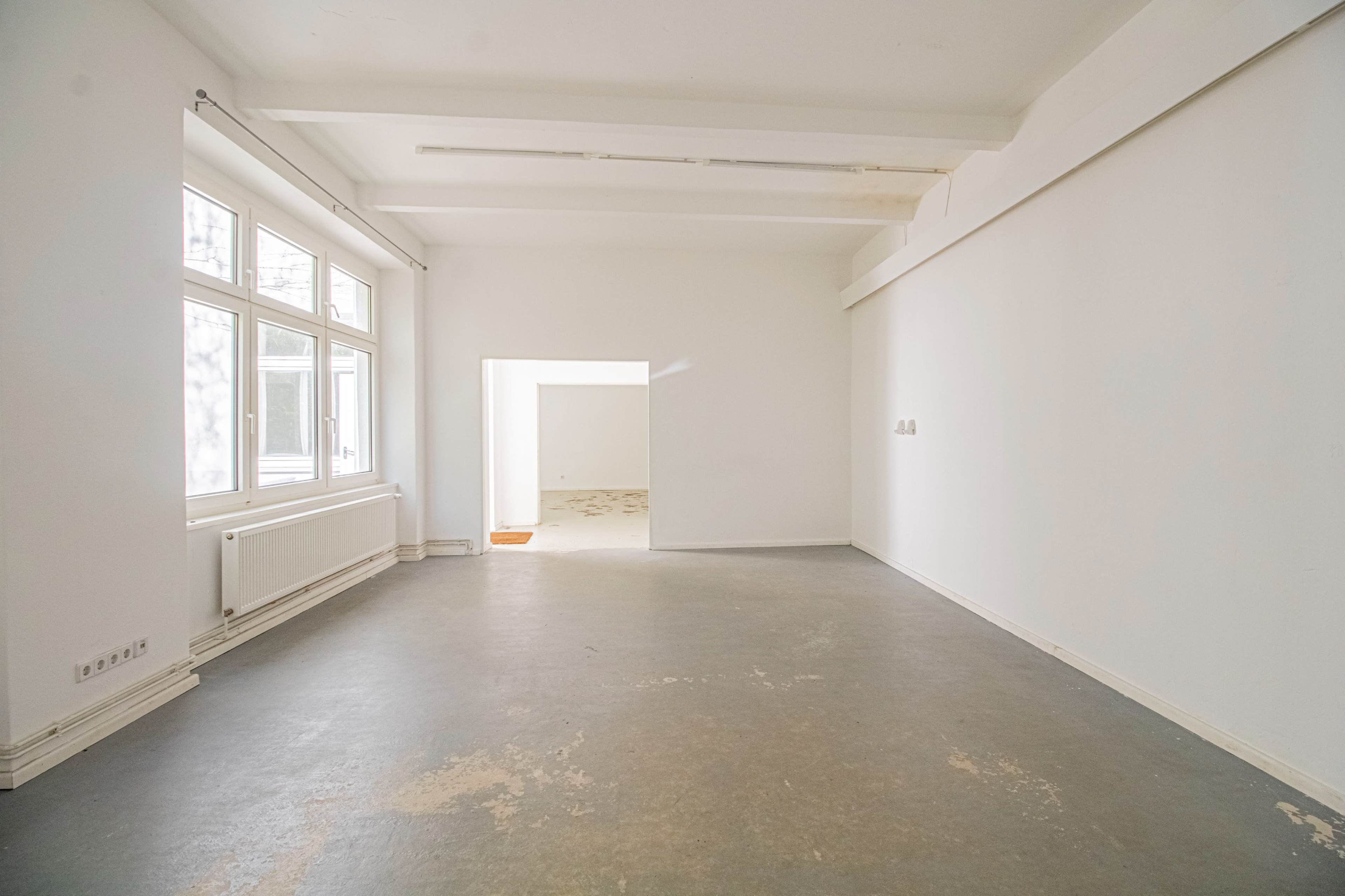 The image shows a sparsely furnished room with large windows and a plain, light-colored interior.
