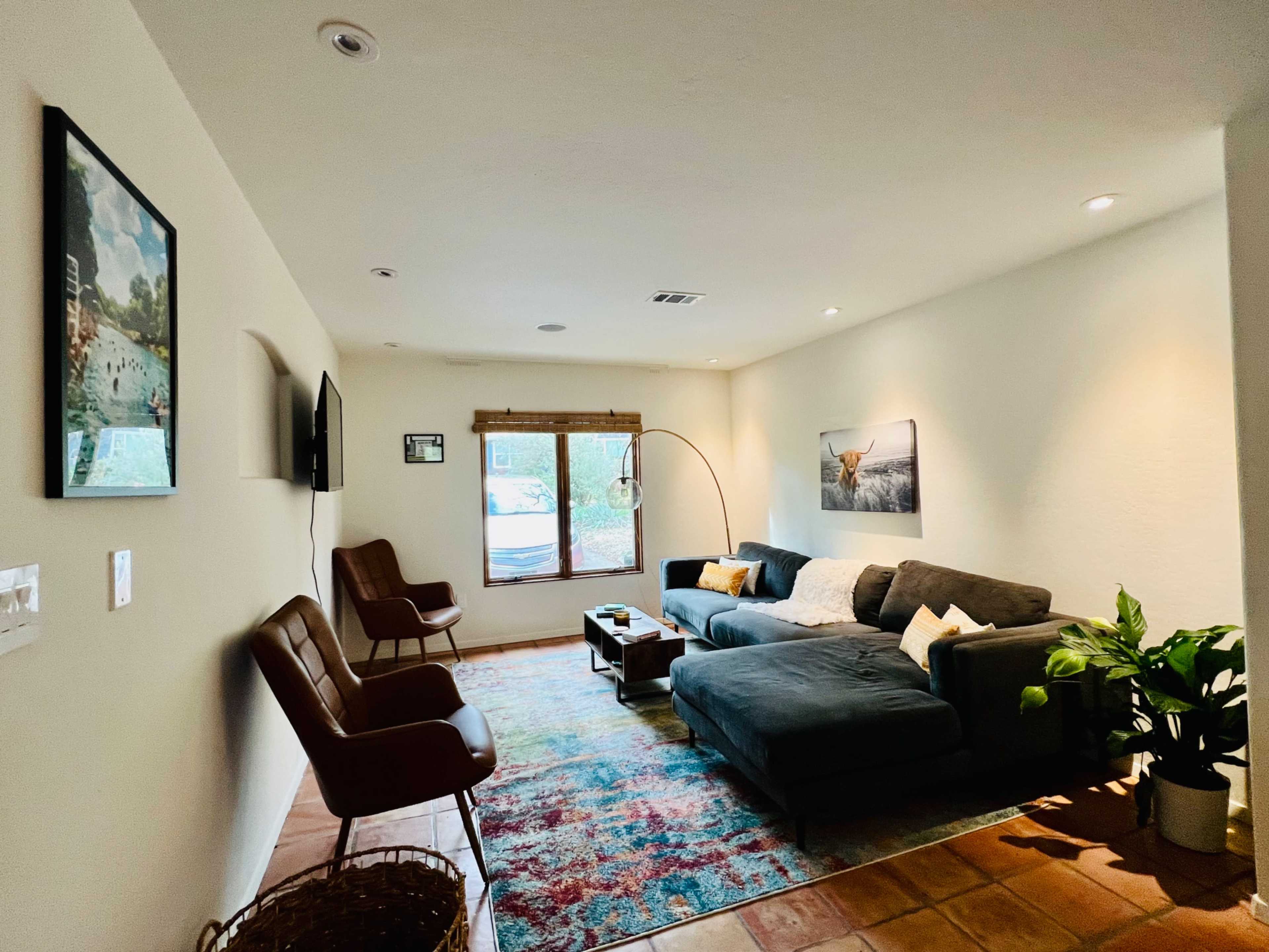 A living room features a gray sectional sofa, two brown chairs, a floor lamp, and a colorful area rug, with wall art and a window letting in natural light.