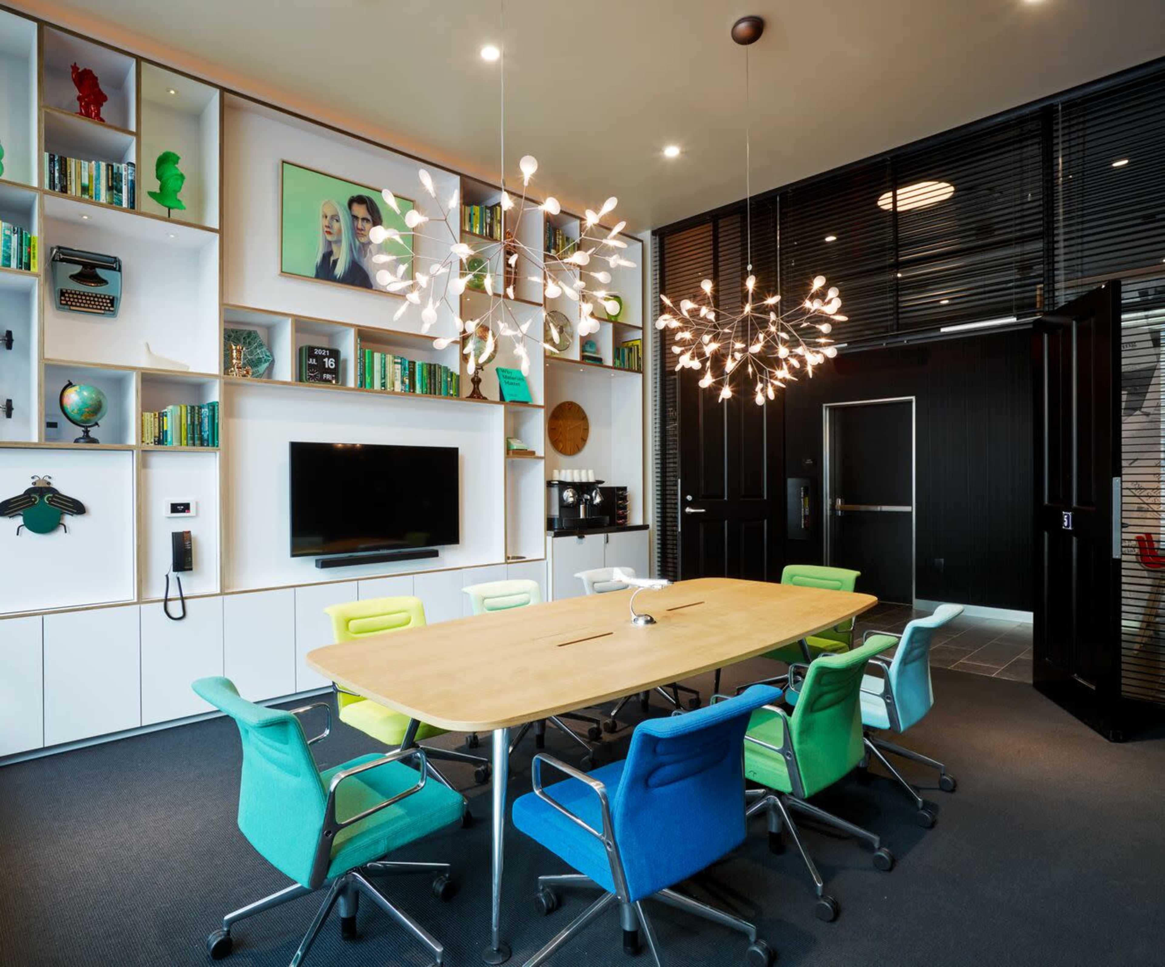 A modern meeting room features a large wooden table surrounded by colorful chairs, with a bookshelf displaying decor and books, and two distinctive light fixtures hanging from the ceiling.
