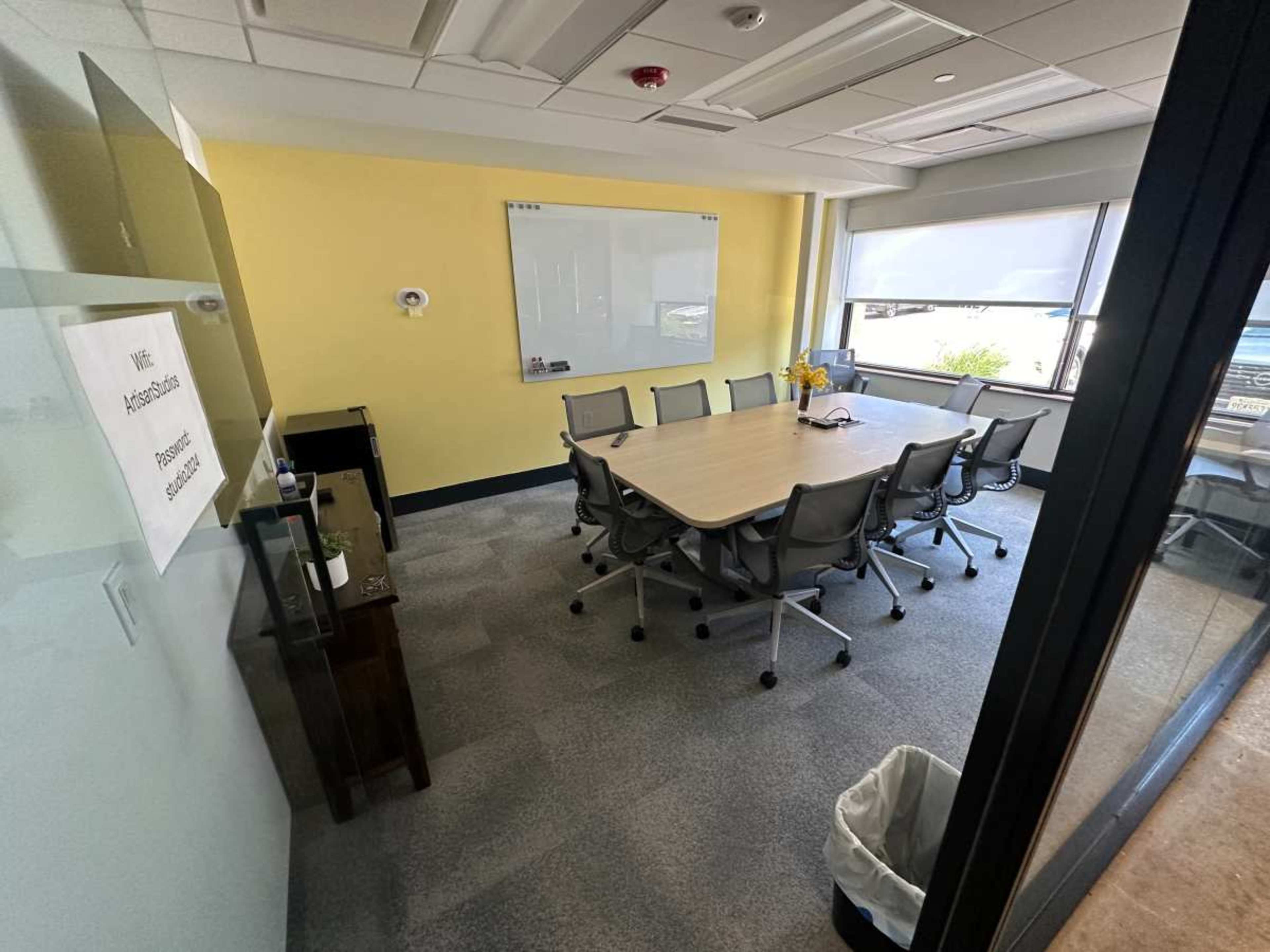 A conference room features a large table surrounded by chairs, a whiteboard on the wall, and a window with blinds.
