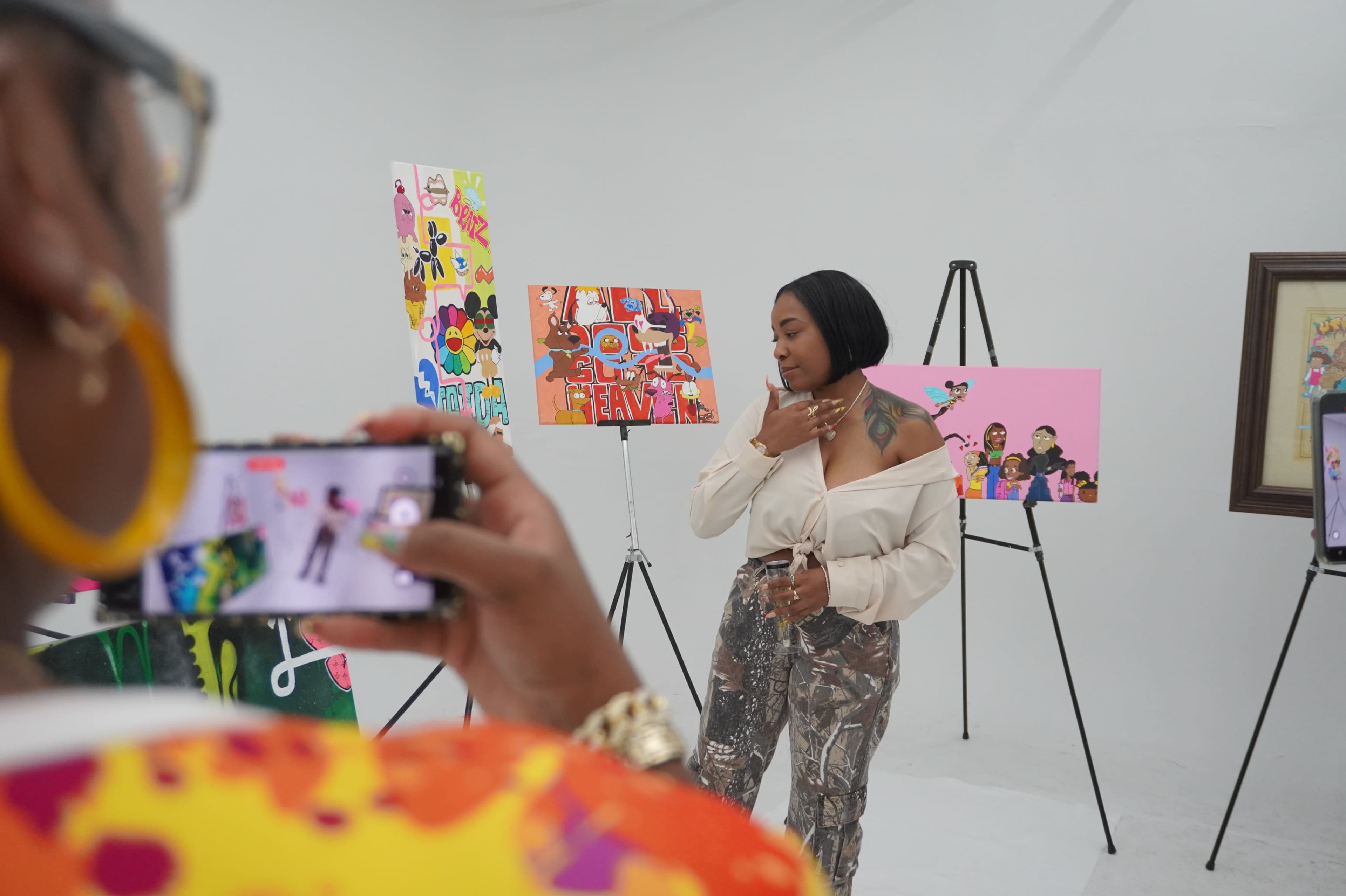 A woman poses in front of colorful artwork while another person captures the moment with a camera.