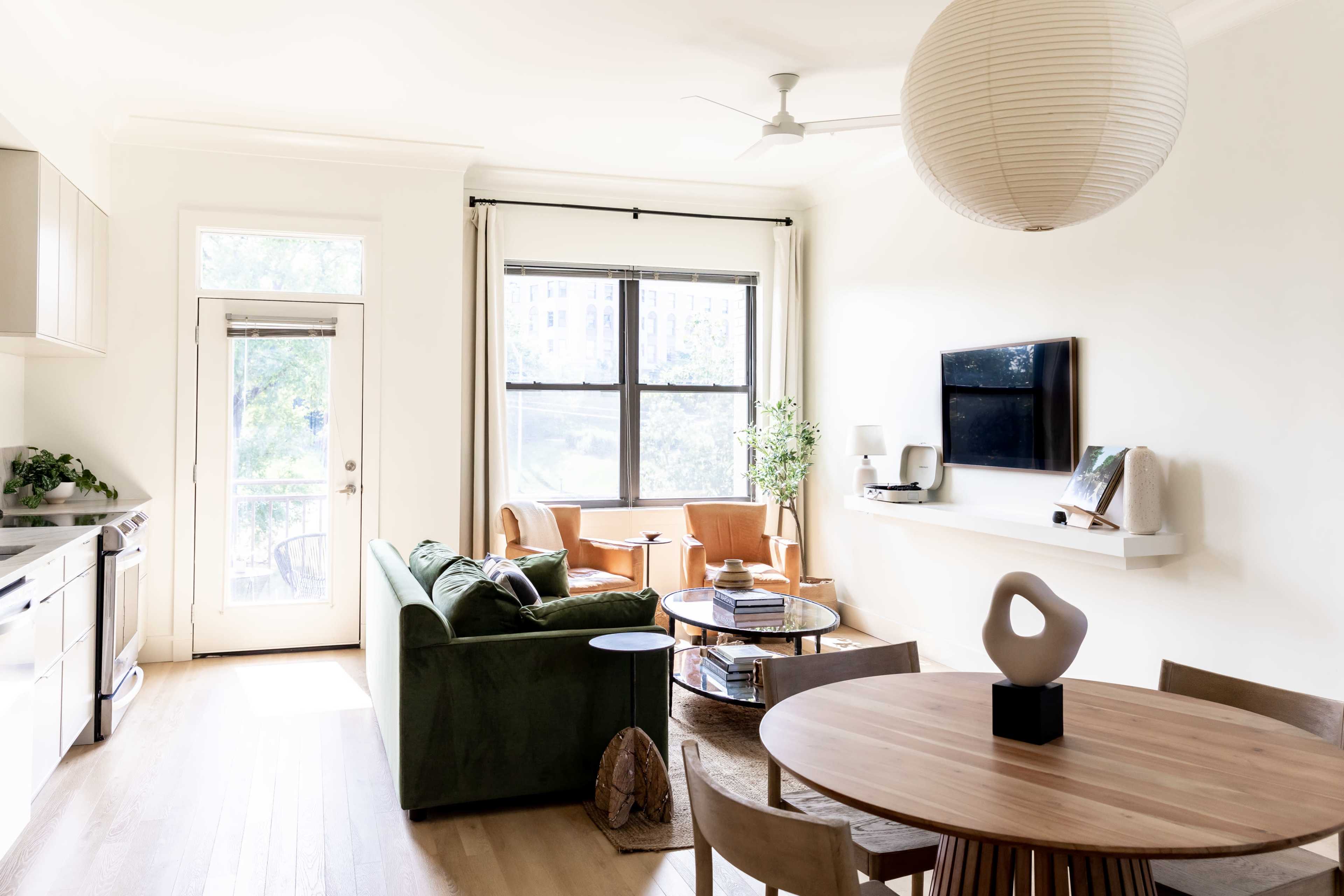A bright living room features a green sofa, a wooden dining table, and a flat-screen TV mounted on the wall, with large windows allowing natural light.