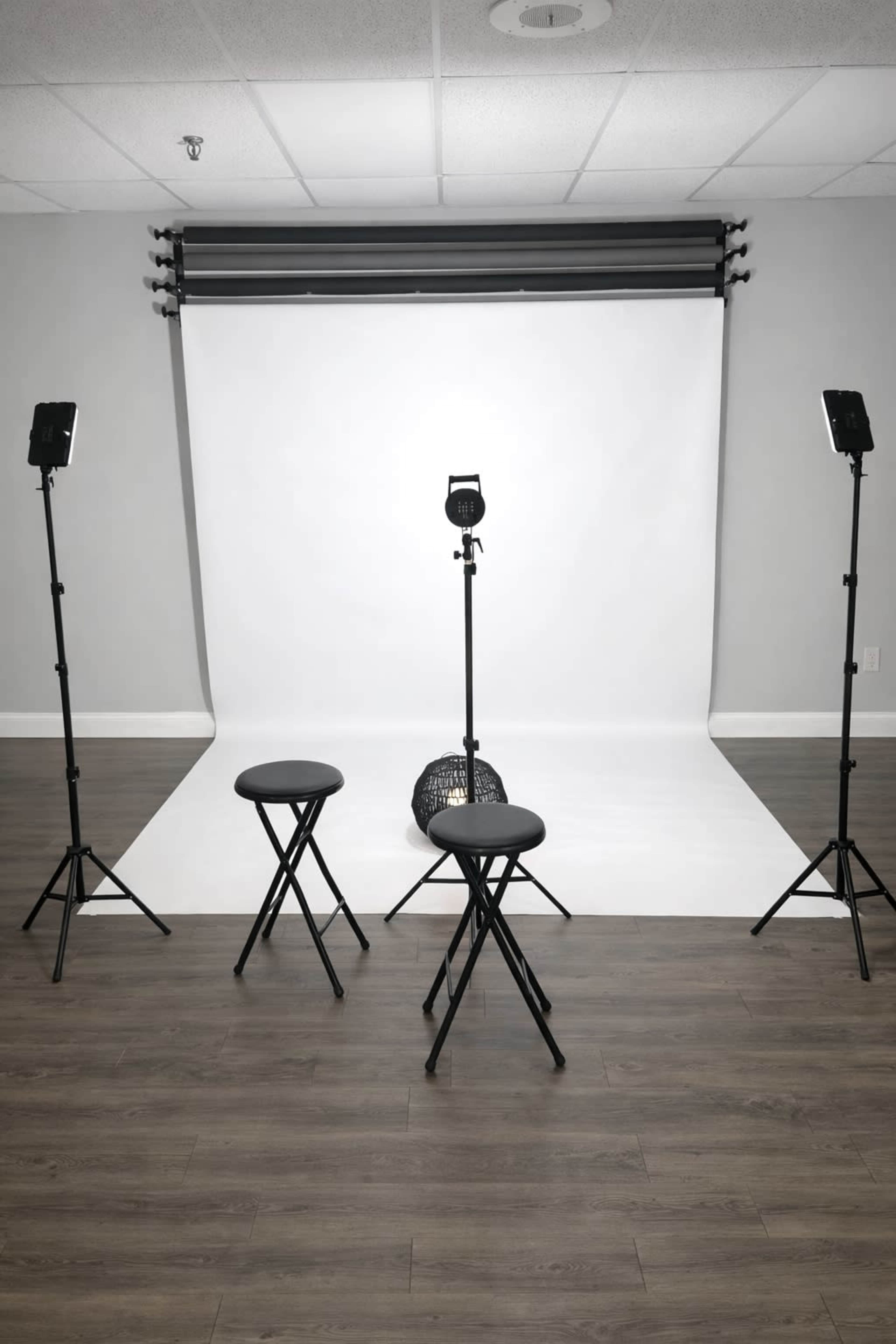 The image shows a photography studio setup with a white backdrop, three black stools, and two lighting stands positioned around the area.