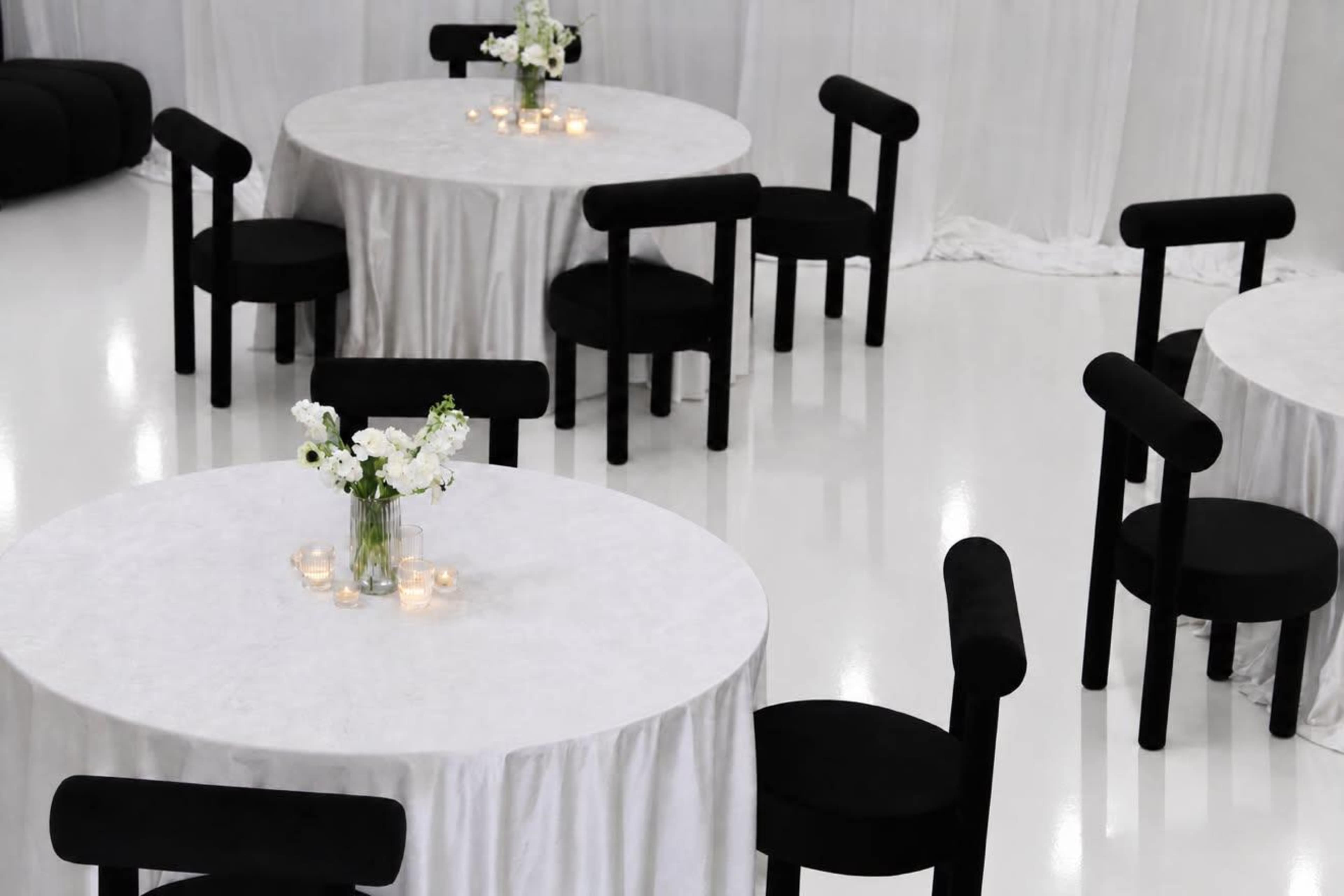 The image shows several round tables covered with white tablecloths, surrounded by black chairs, and decorated with small flower arrangements and candles.