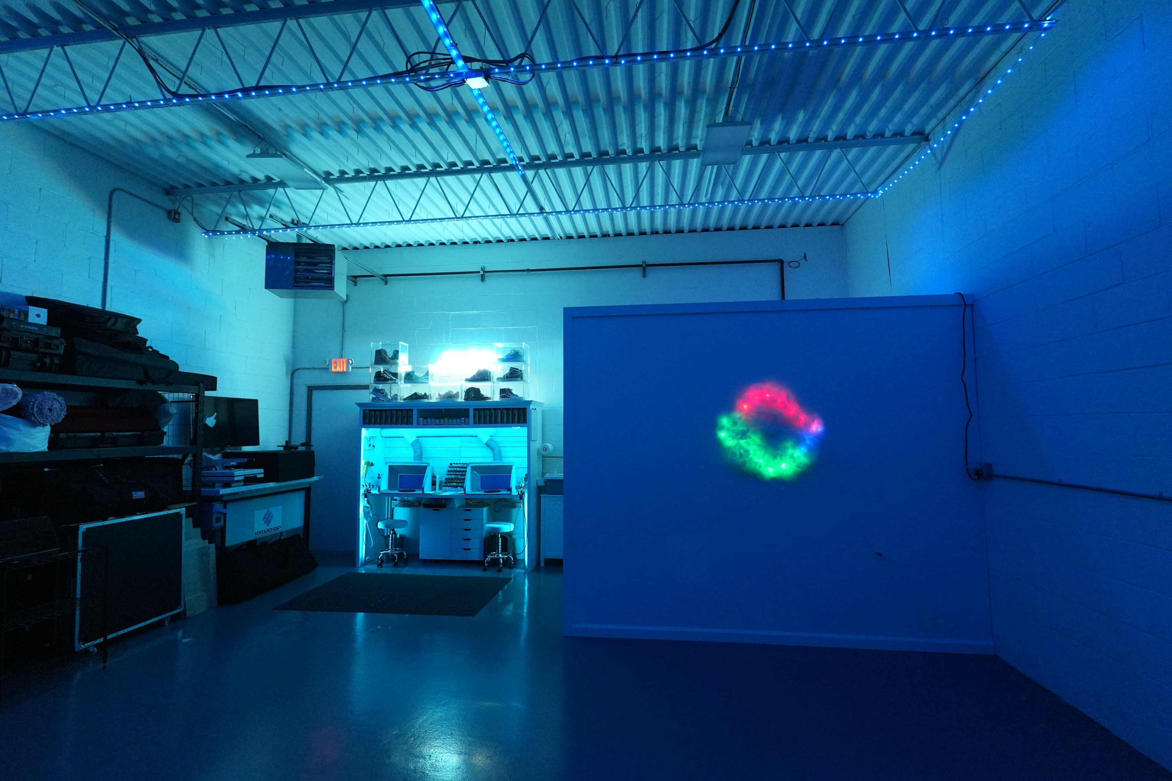 The image shows a dimly lit room with blue lighting, featuring a wall that displays a colorful, illuminated pattern and various equipment in the background.