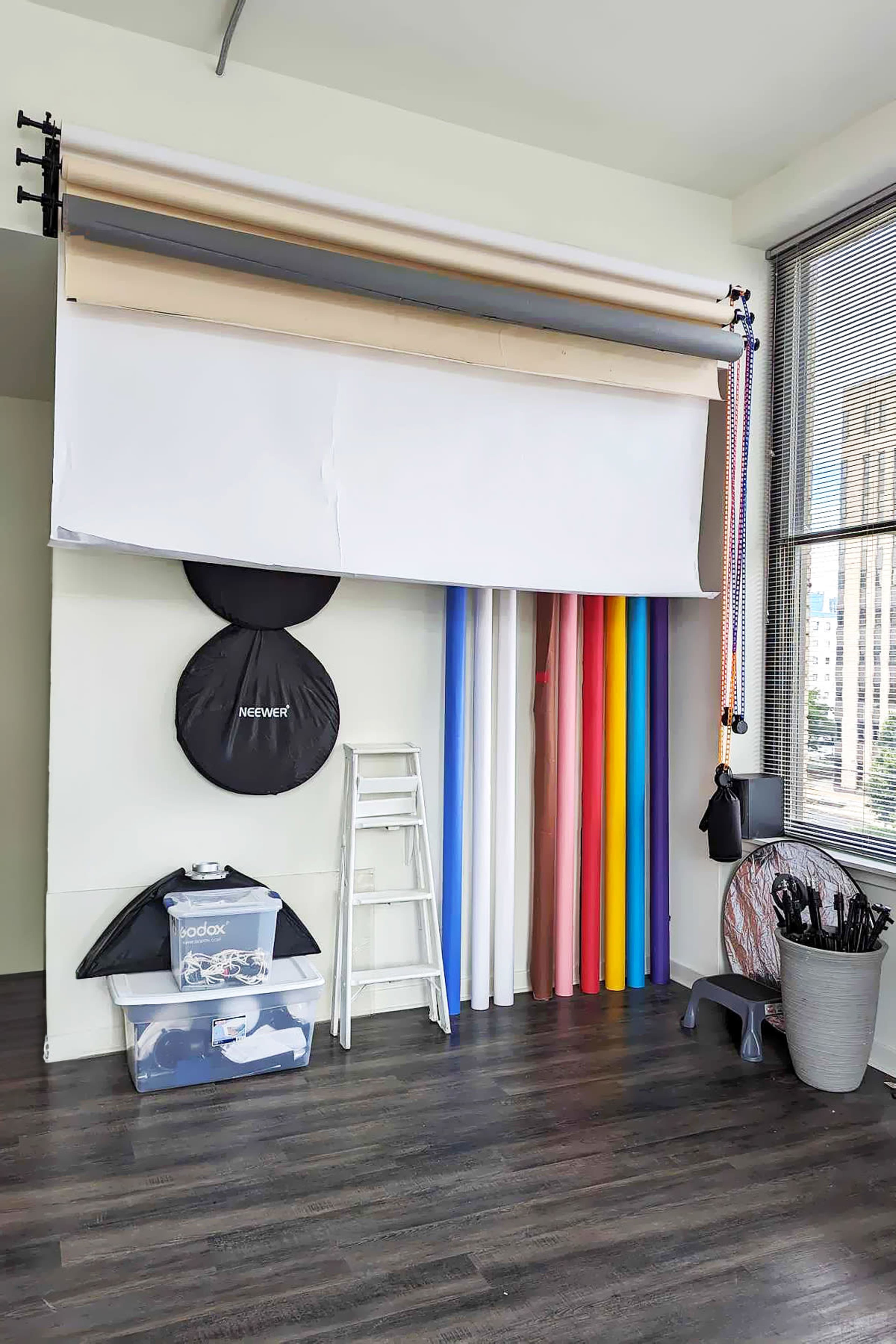 A photography studio with a backdrop roller system and various colored backdrop paper rolls neatly arranged beside a ladder and storage containers.