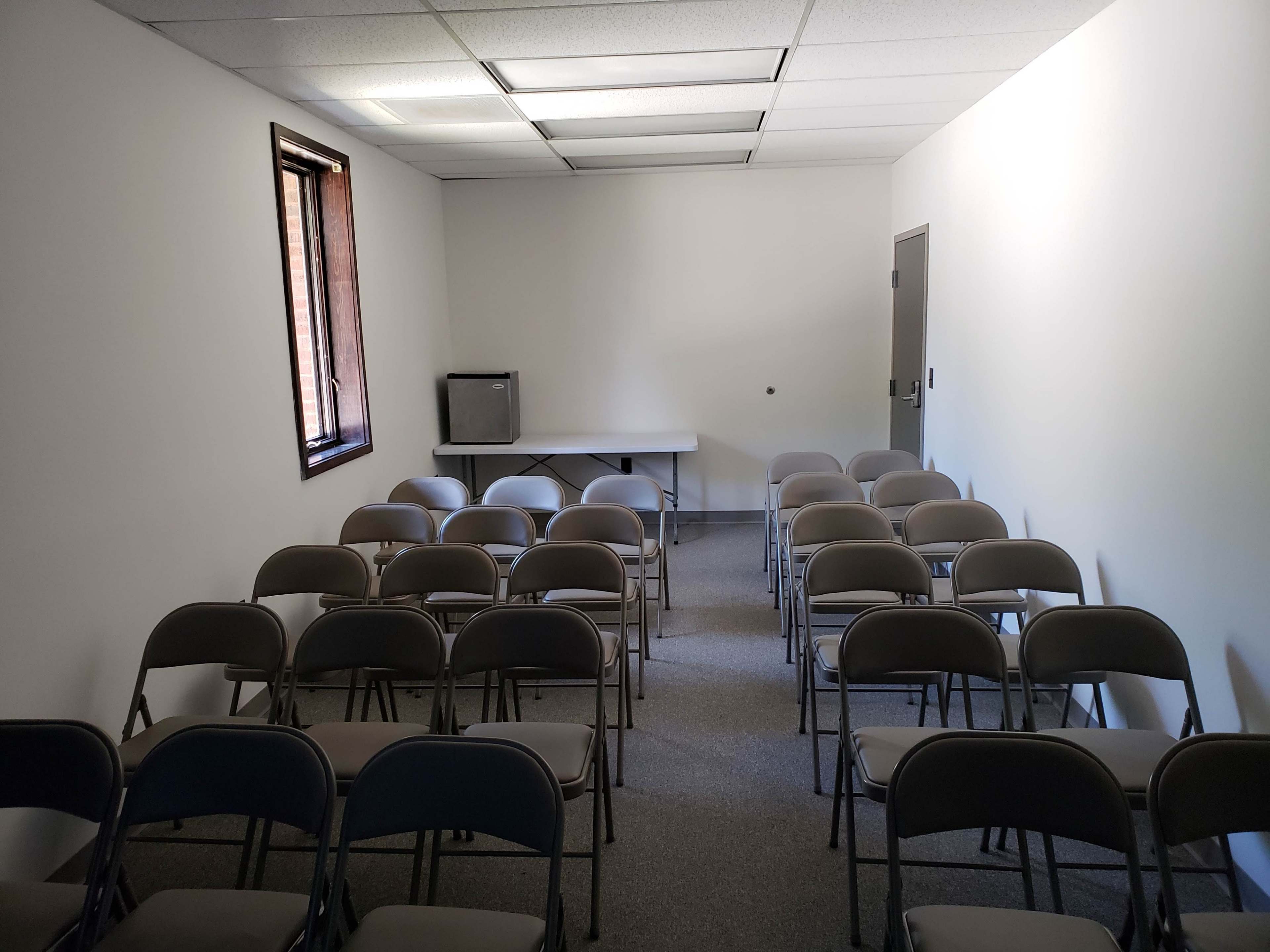 The image shows a small, empty room arranged with rows of folding chairs facing a table, with a window and a television in the corner.