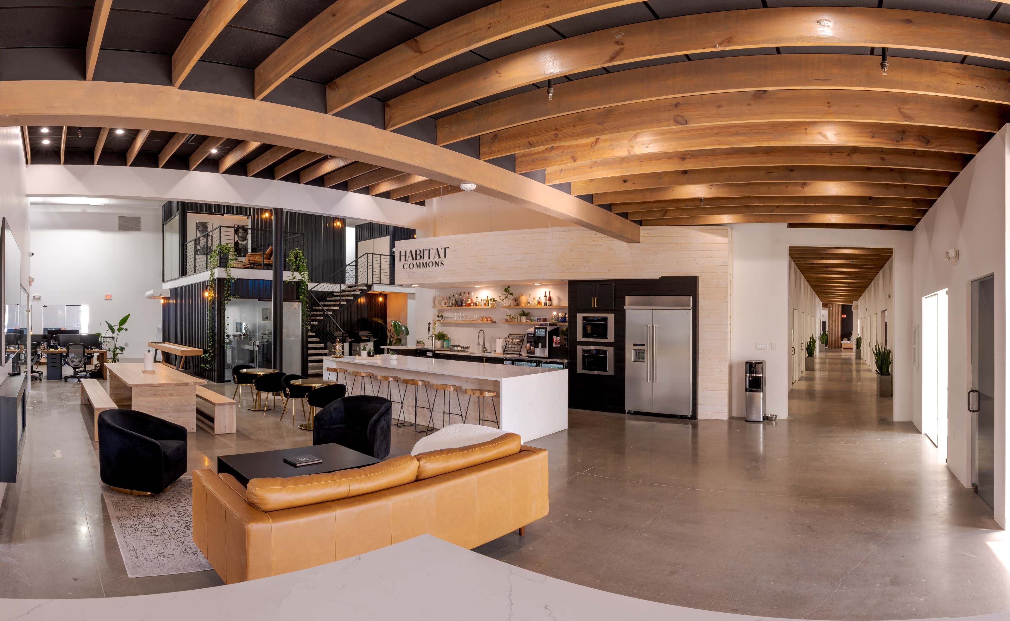 The image shows a modern office space featuring an open layout with exposed wooden beams, a kitchen area with seating, and a corridor leading to other sections.