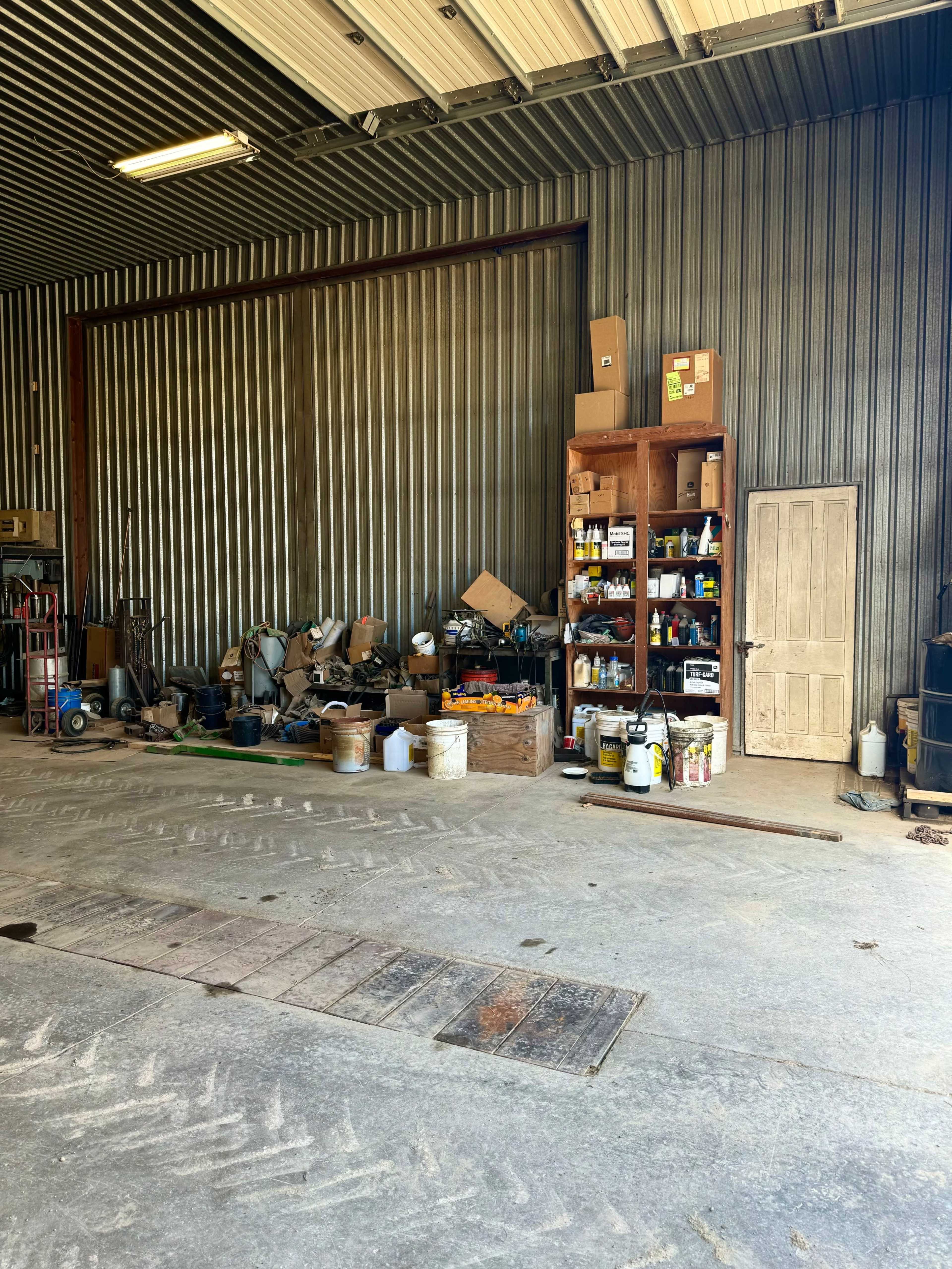 The image shows the interior of a workshop with a metal-clad wall, a wooden shelf filled with various tools and supplies, and a door on the far side.