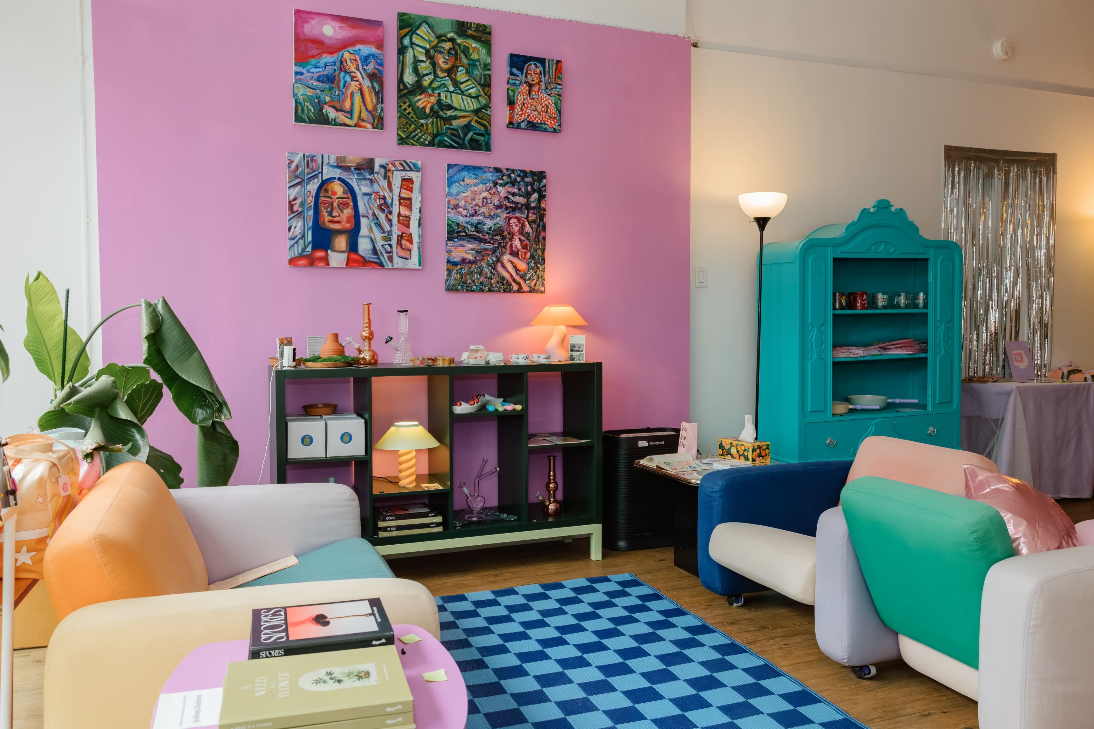 The image shows a colorful living room with pastel-colored furniture, a blue checkered rug, and artwork displayed on a pink wall.