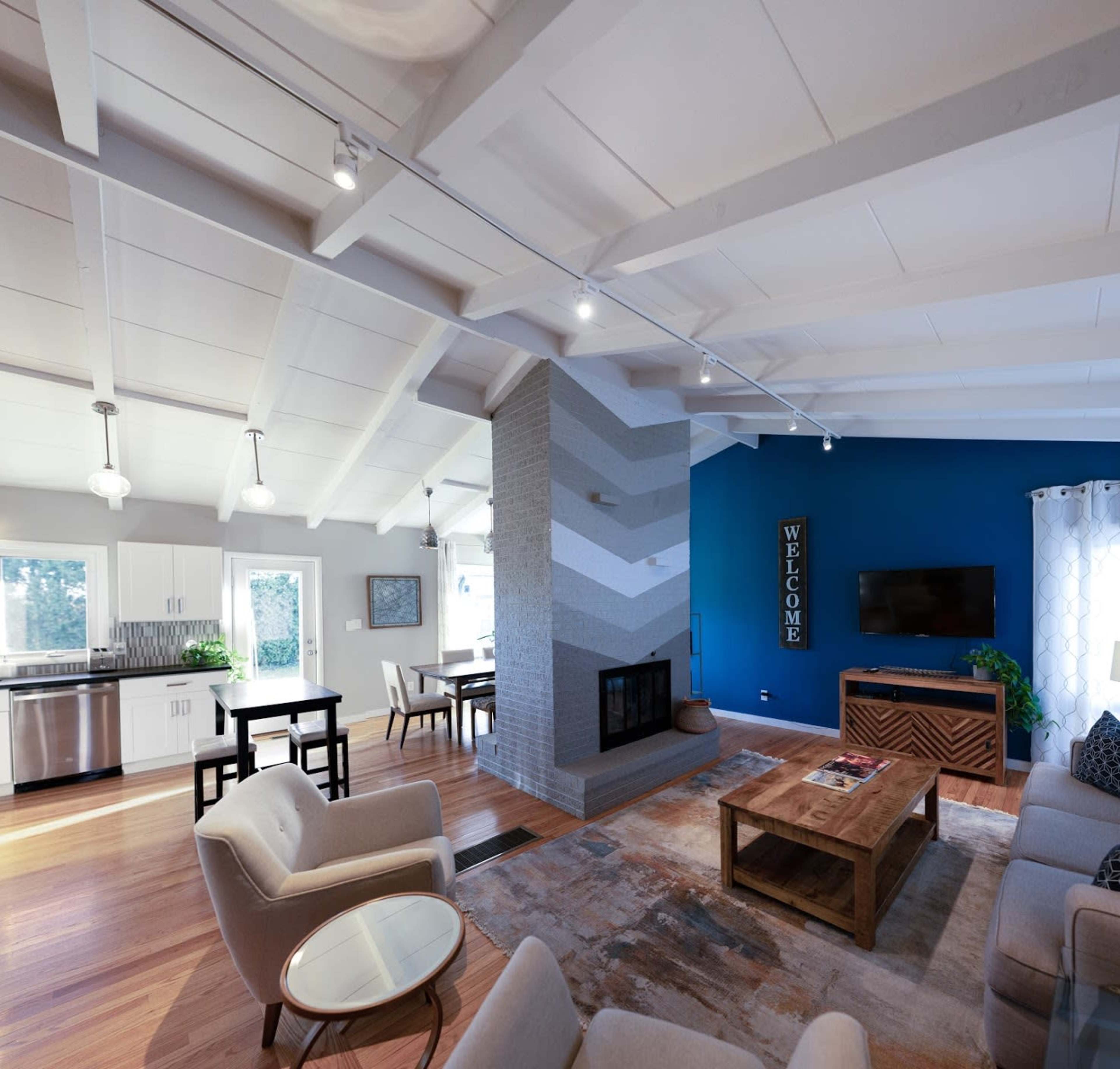 A modern living room with light wood floors, a central fireplace, and a blue accent wall, featuring a dining area and kitchen in the background.