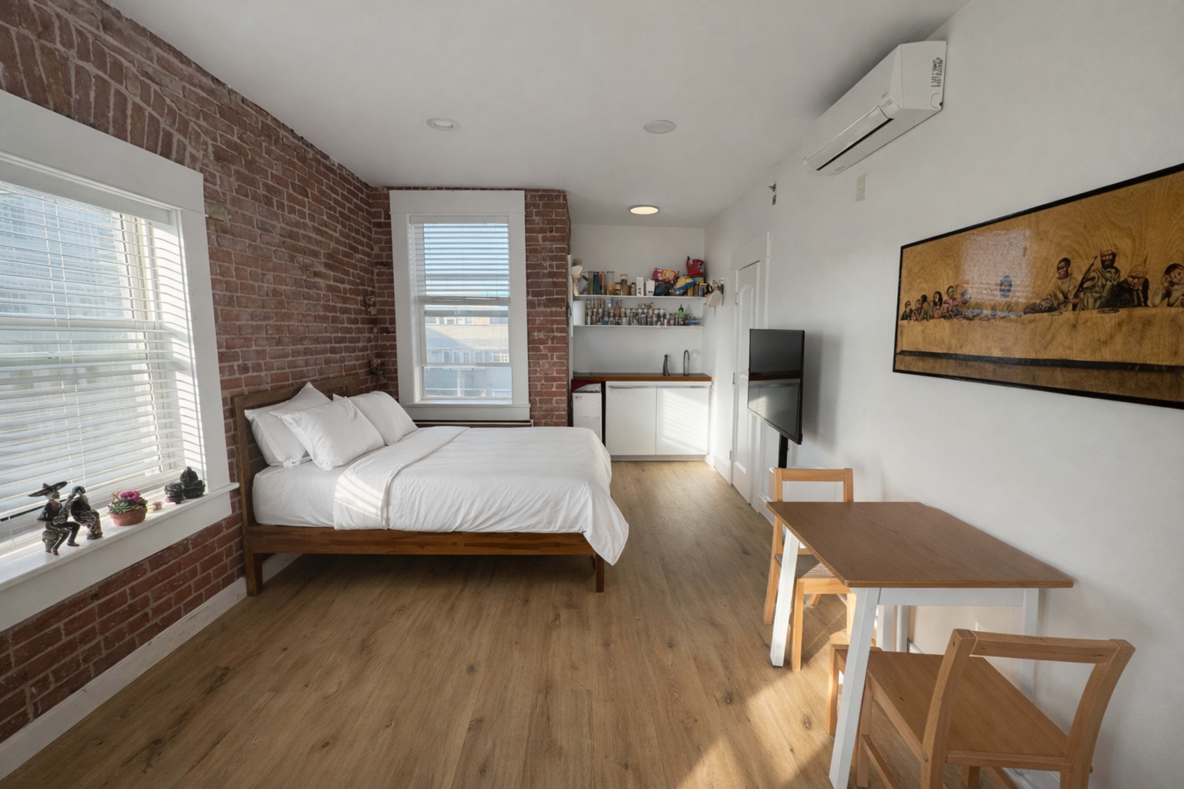 The image depicts a small bedroom with a bed, a table and chairs, a television, and exposed brick walls.