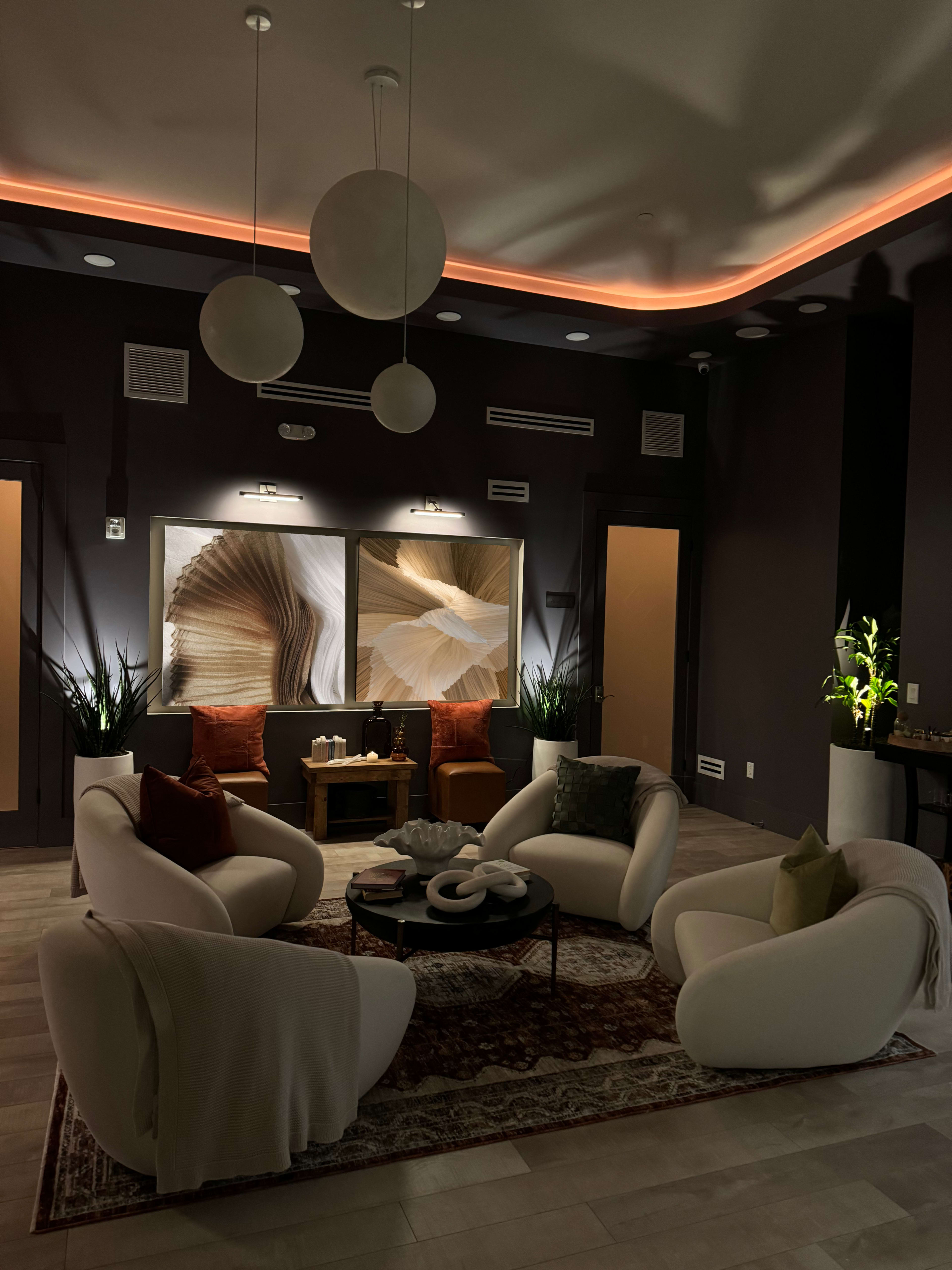A cozy, modern lounge features four white chairs arranged around a circular black table, with warm lighting highlighting two large artwork pieces on the wall.