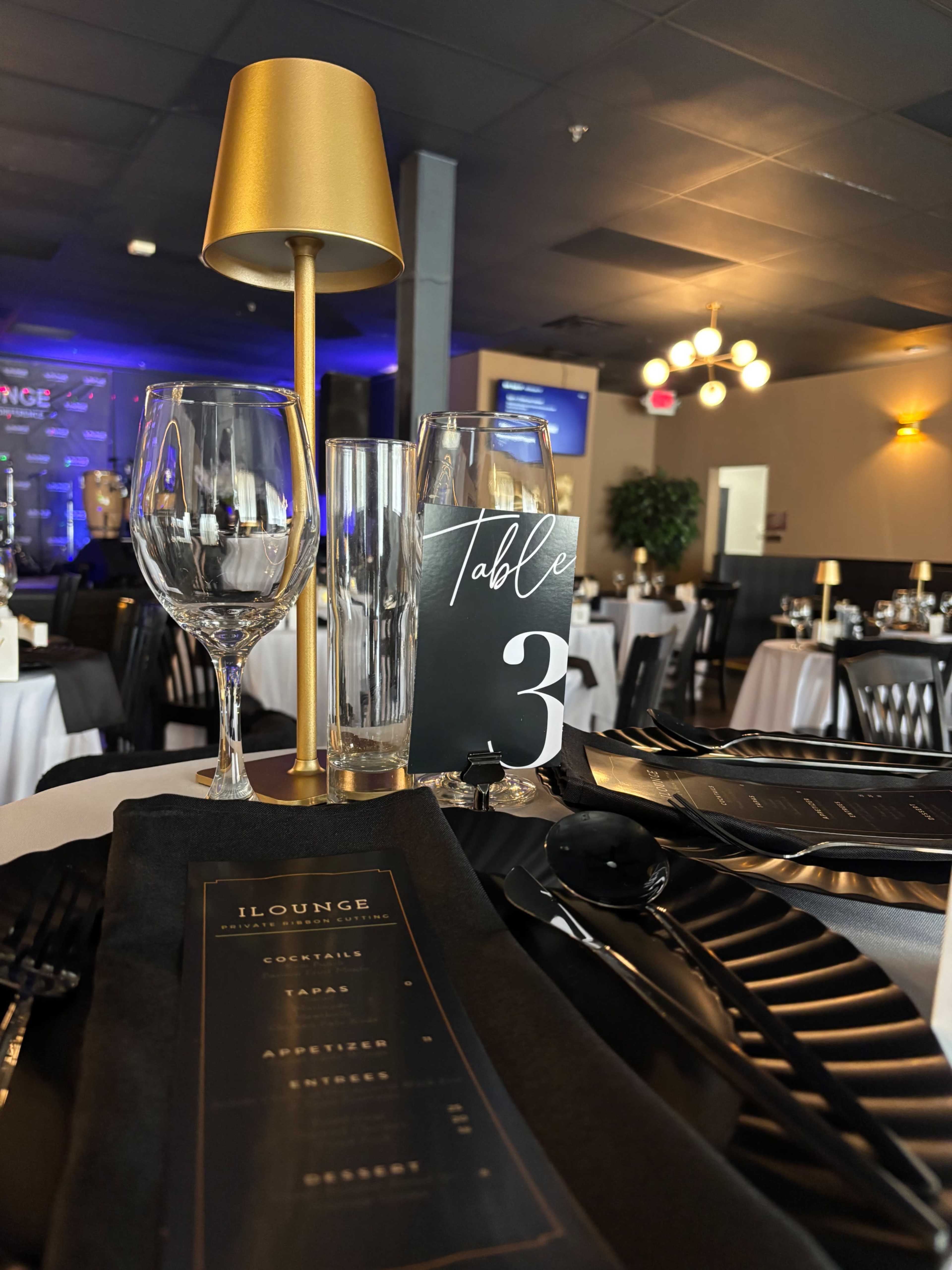 The image shows a neatly set dining table with a gold lamp, elegant glassware, a black table tent indicating "Table 3," and a black menu laid out on a white tablecloth.