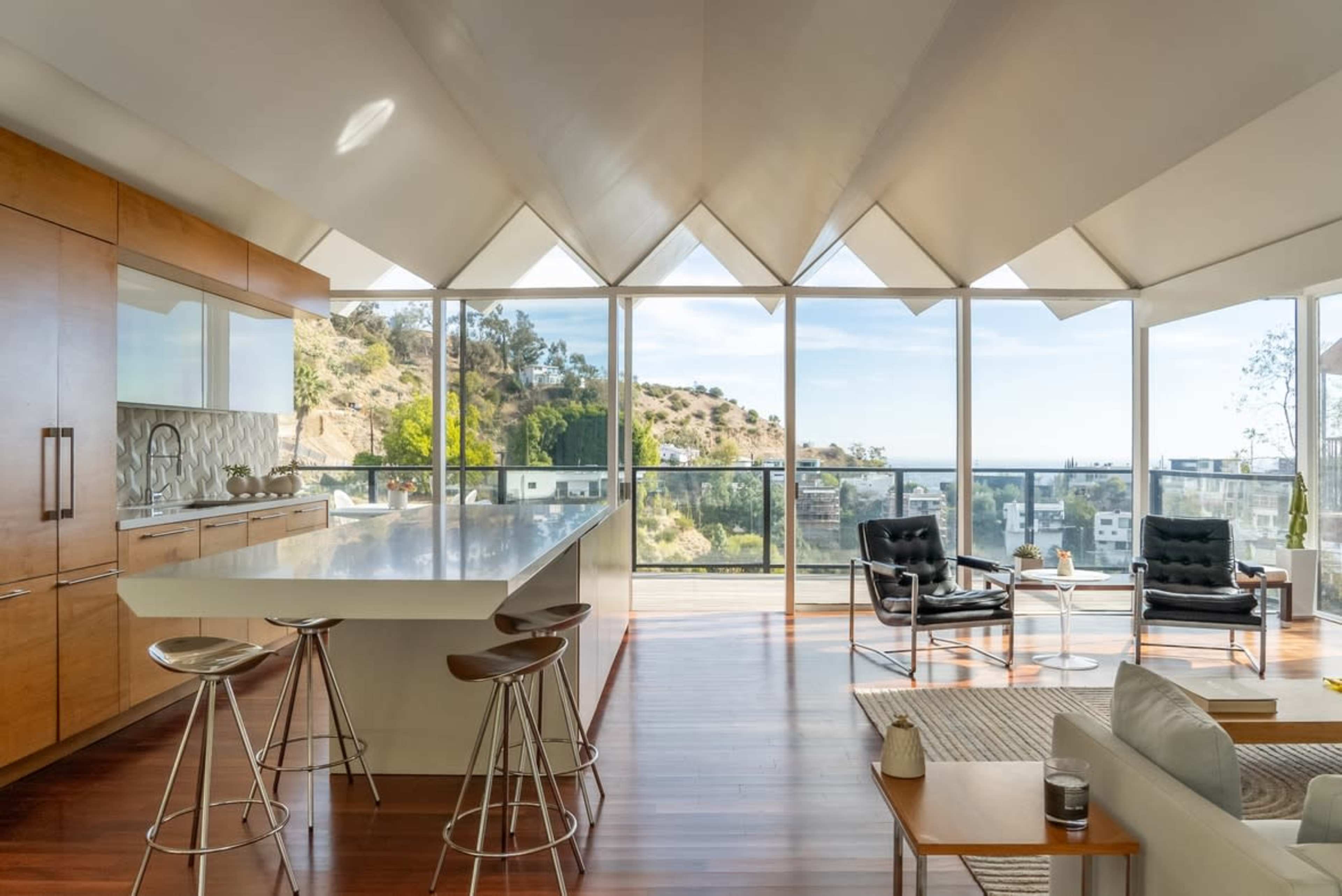 The image shows a modern, open-concept living space with a large kitchen island, floor-to-ceiling windows, and a view of a hillside outside.