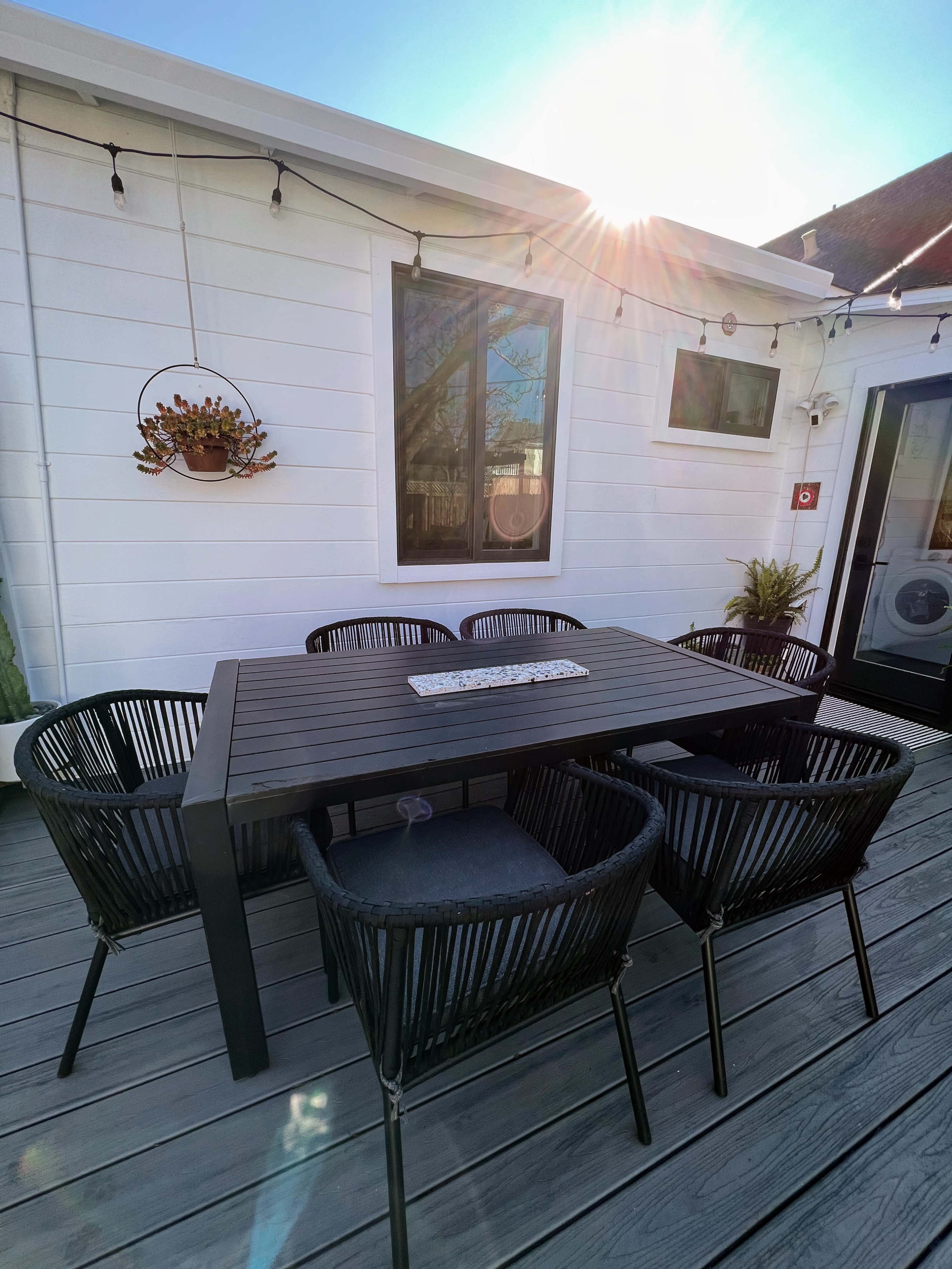 A wooden dining table with six black woven chairs is set on a deck beside a white house.