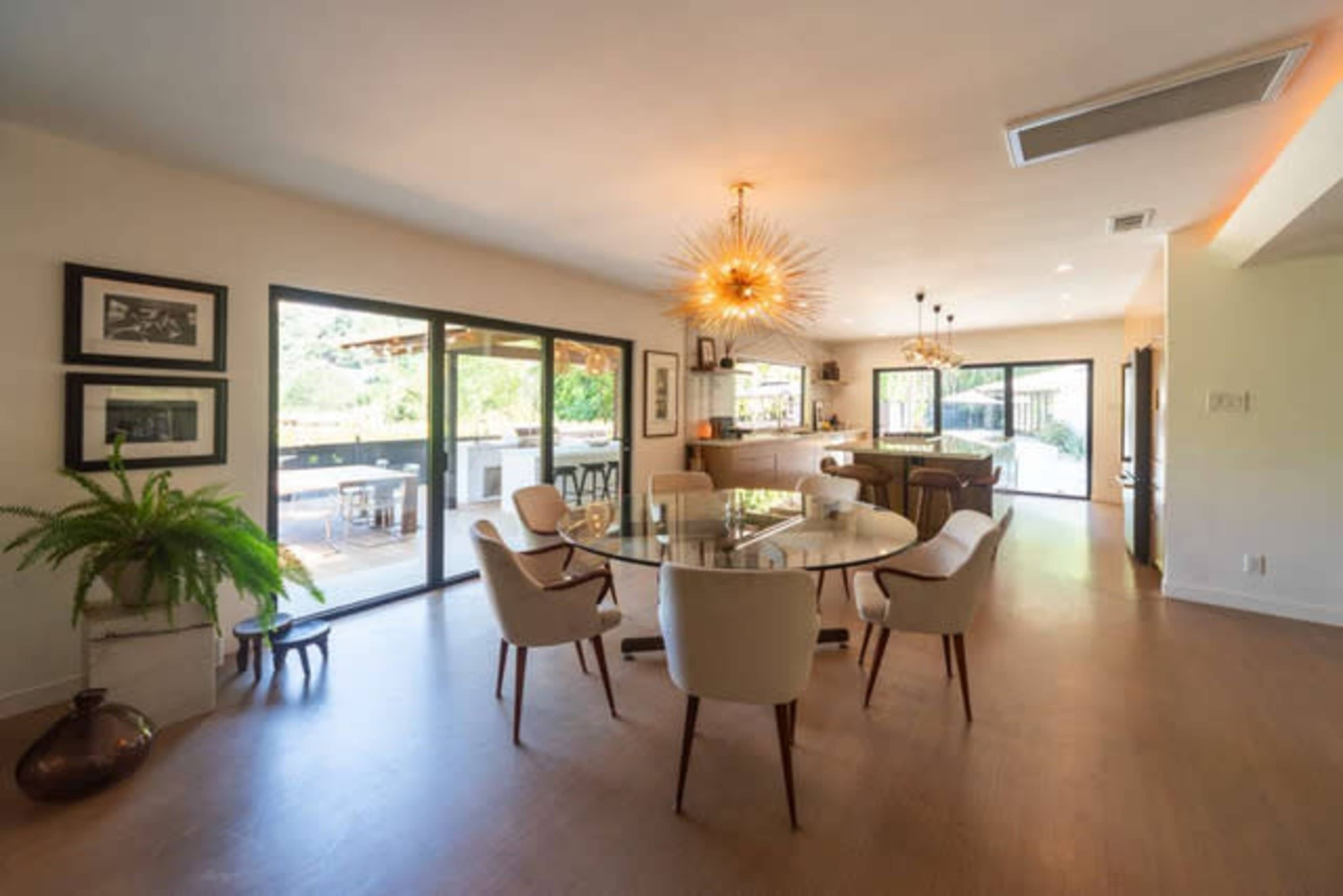 The dining area features a round glass table surrounded by upholstered chairs, with large windows overlooking an outdoor space.
