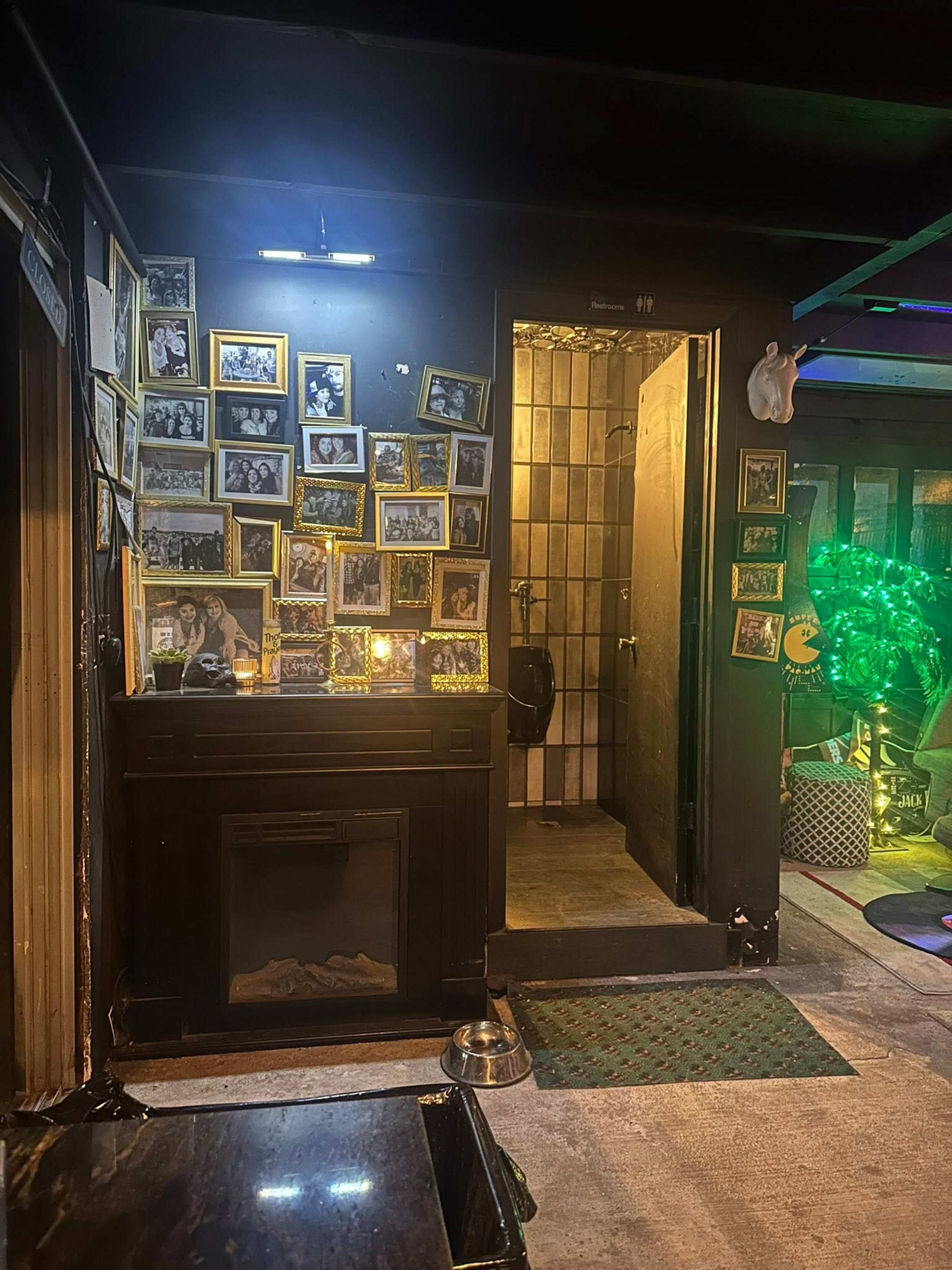 The image shows a dimly lit entrance to a bar or restaurant, featuring a wall adorned with framed photographs and a restroom door on the right.