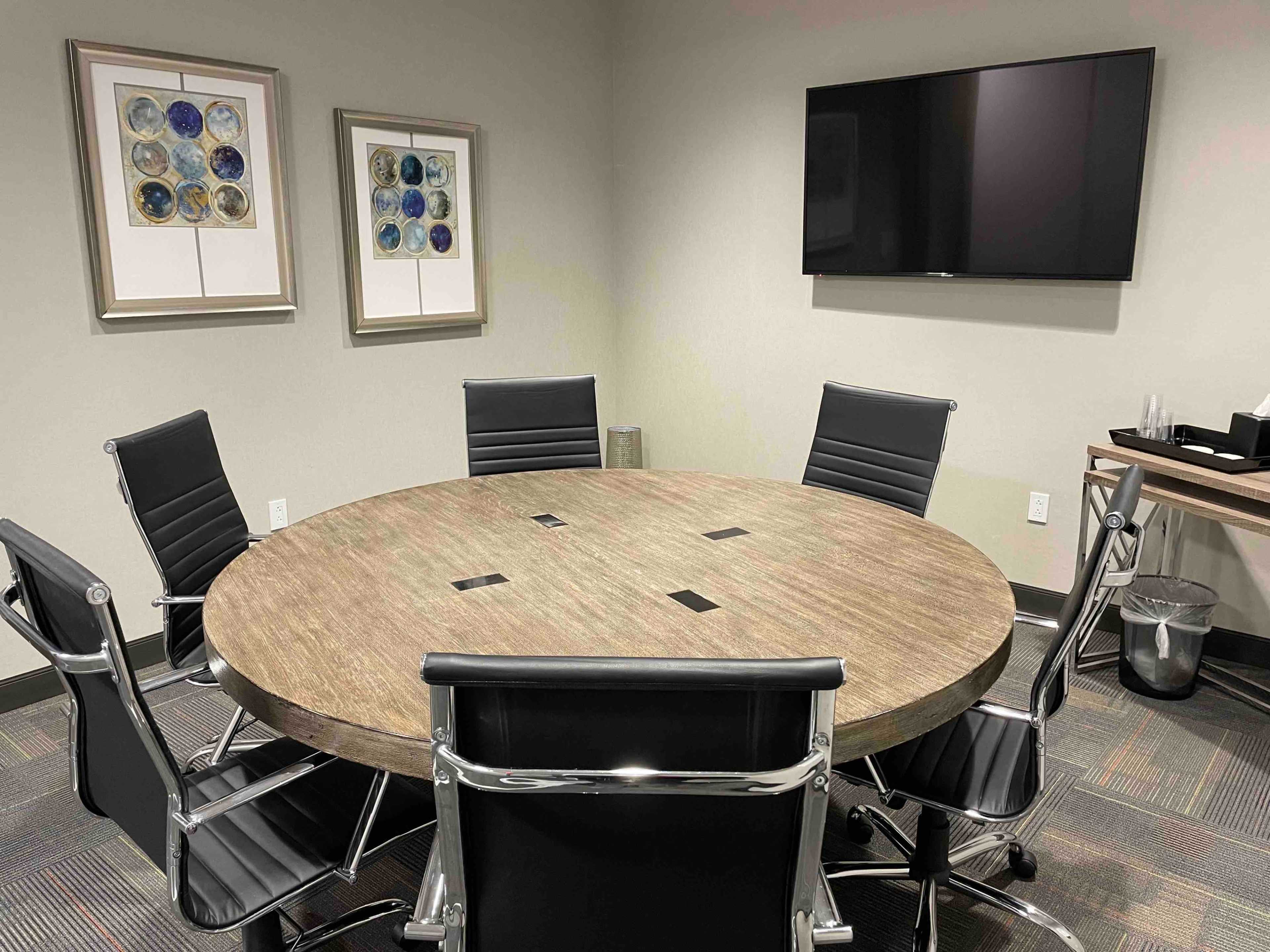 A round wooden conference table is surrounded by six black leather chairs in a meeting room with framed artwork and a wall-mounted television.