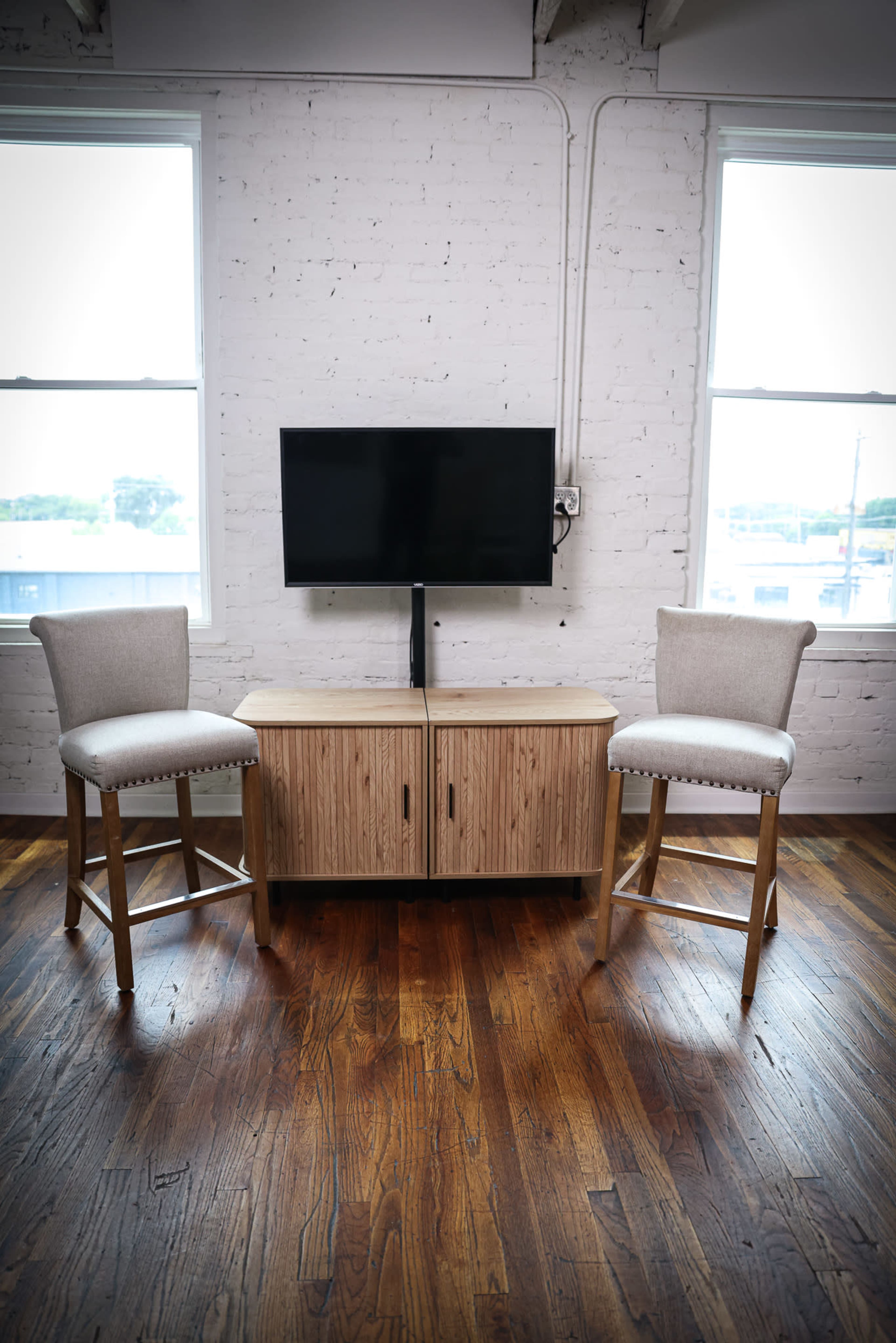 The image shows a modern living space with a wooden cabinet and two barstools positioned in front of a wall-mounted television, all set against a backdrop of large windows and a wooden floor.