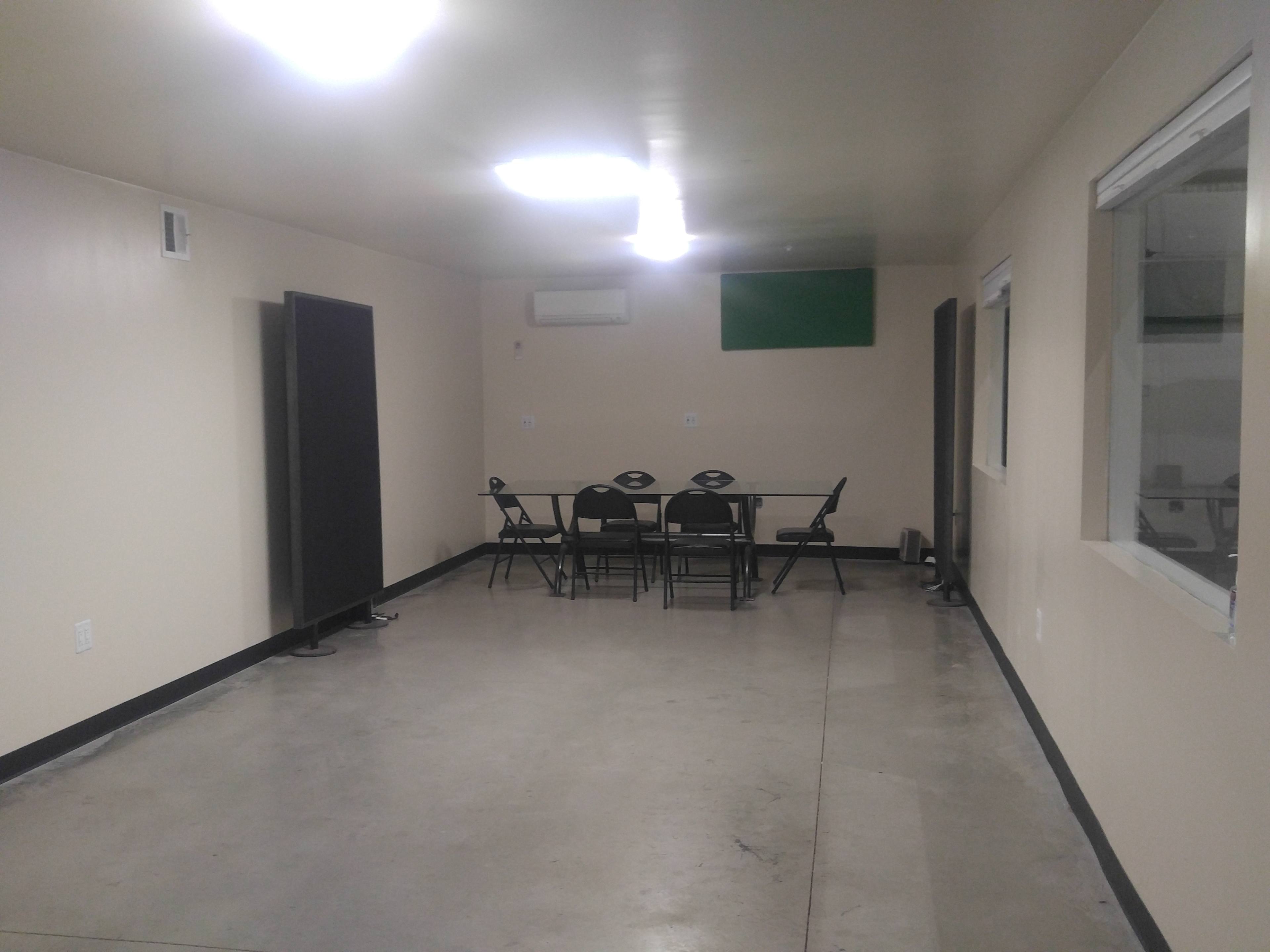 The image shows a sparsely furnished room with a table and chairs at the center, surrounded by plain walls and a concrete floor.