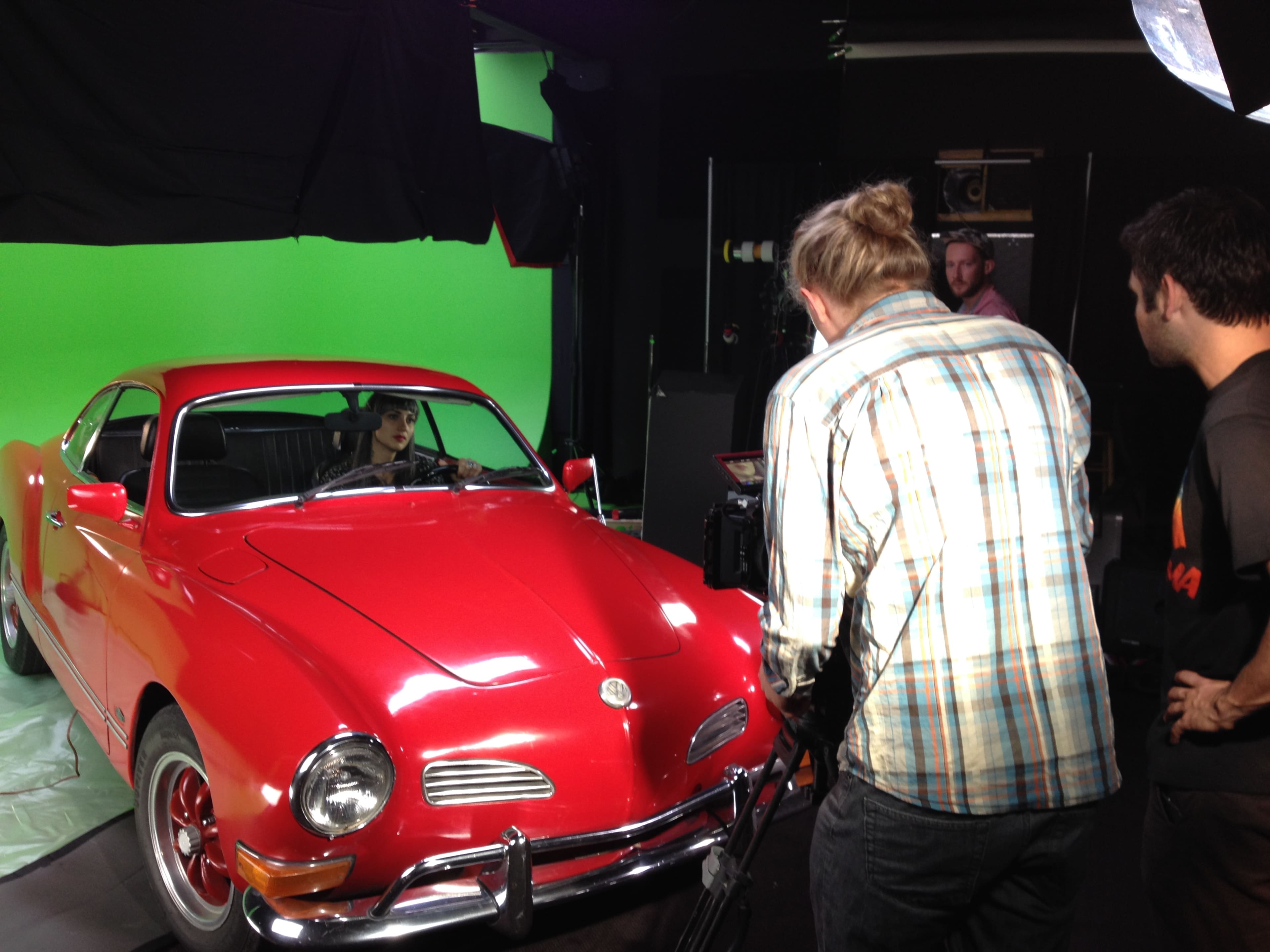 A film crew is filming a scene with a red Volkswagen Karmann Ghia in front of a green screen.