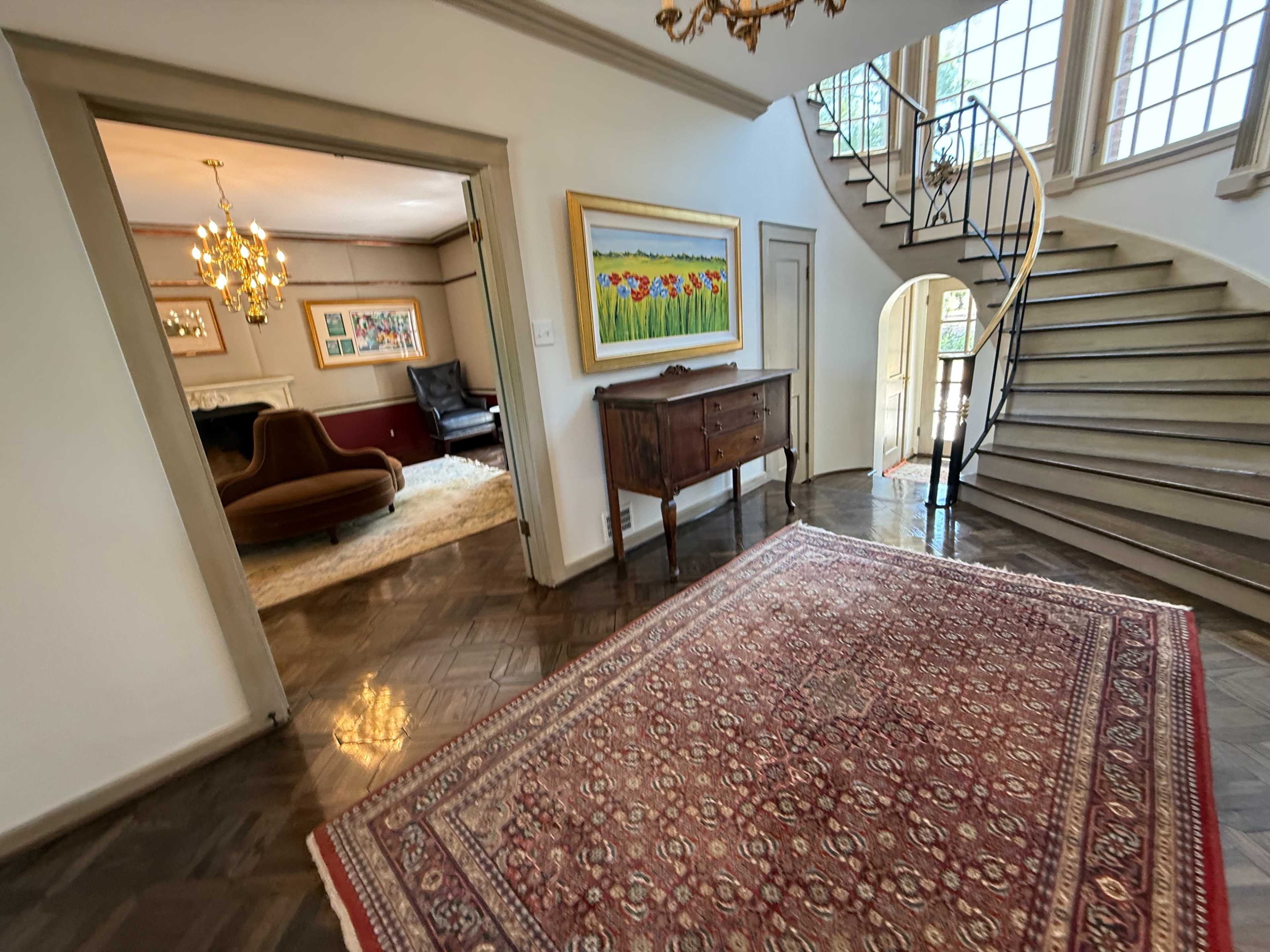The image shows an entryway with a decorative rug, a wooden console table, and a staircase leading to an upper level, with a glimpse of a living area visible through an archway.