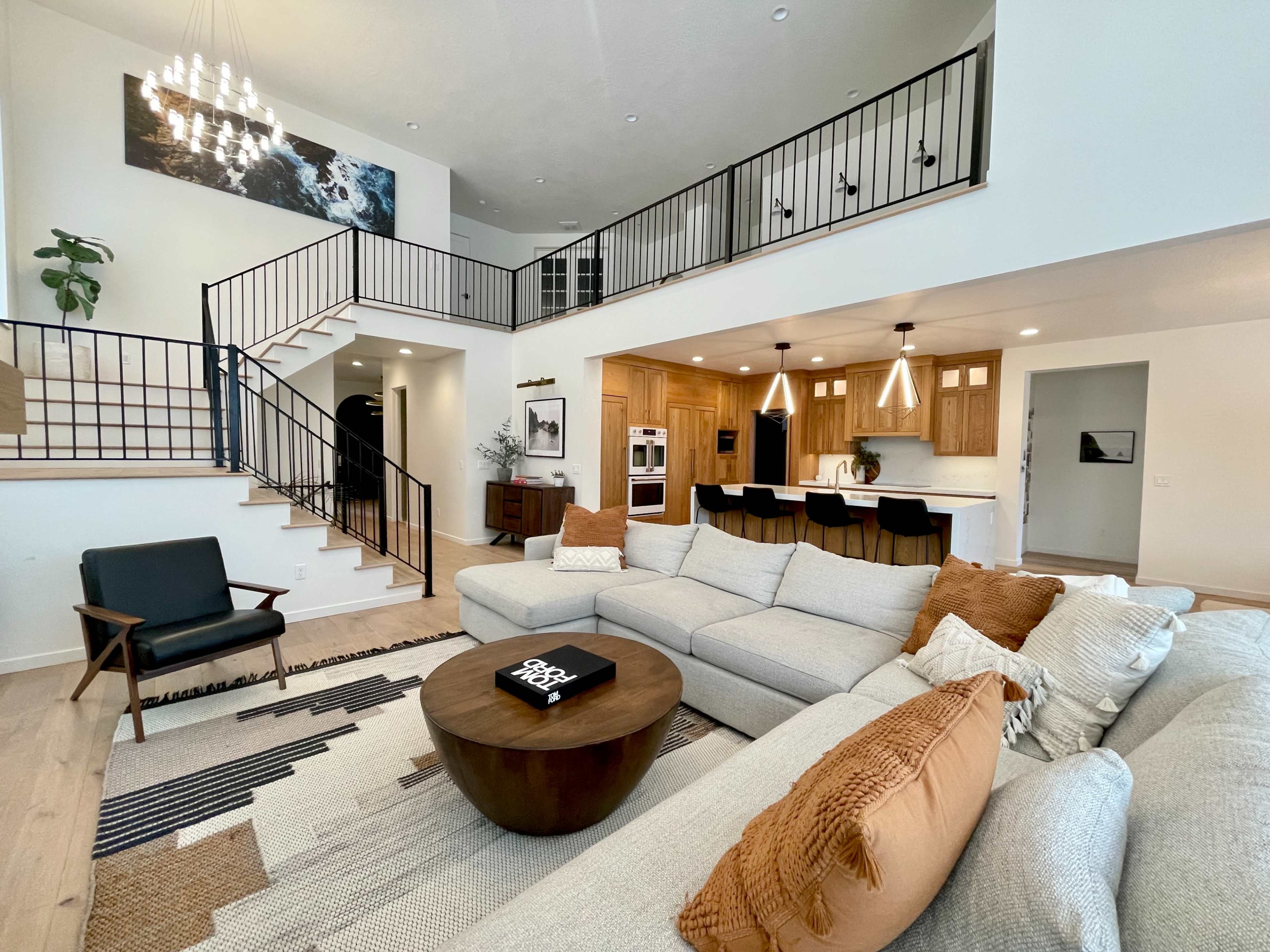 The image shows a modern living room with a sectional sofa, a round coffee table, and a staircase leading to an upper floor, accompanied by a kitchen area featuring wooden cabinets and bar stools.