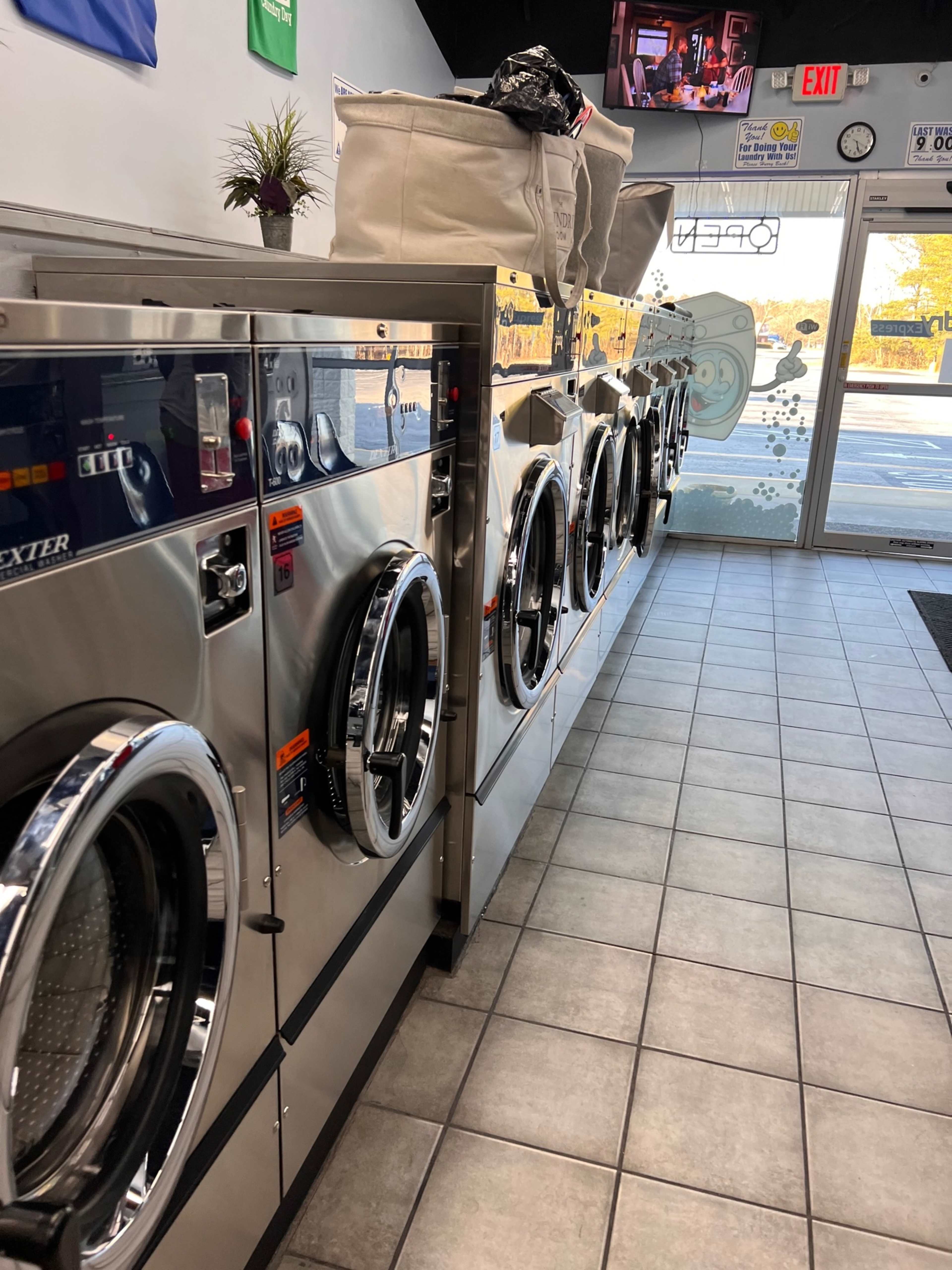 A row of shiny, front-loading washing machines inside a laundromat with large windows and a clear entrance.
