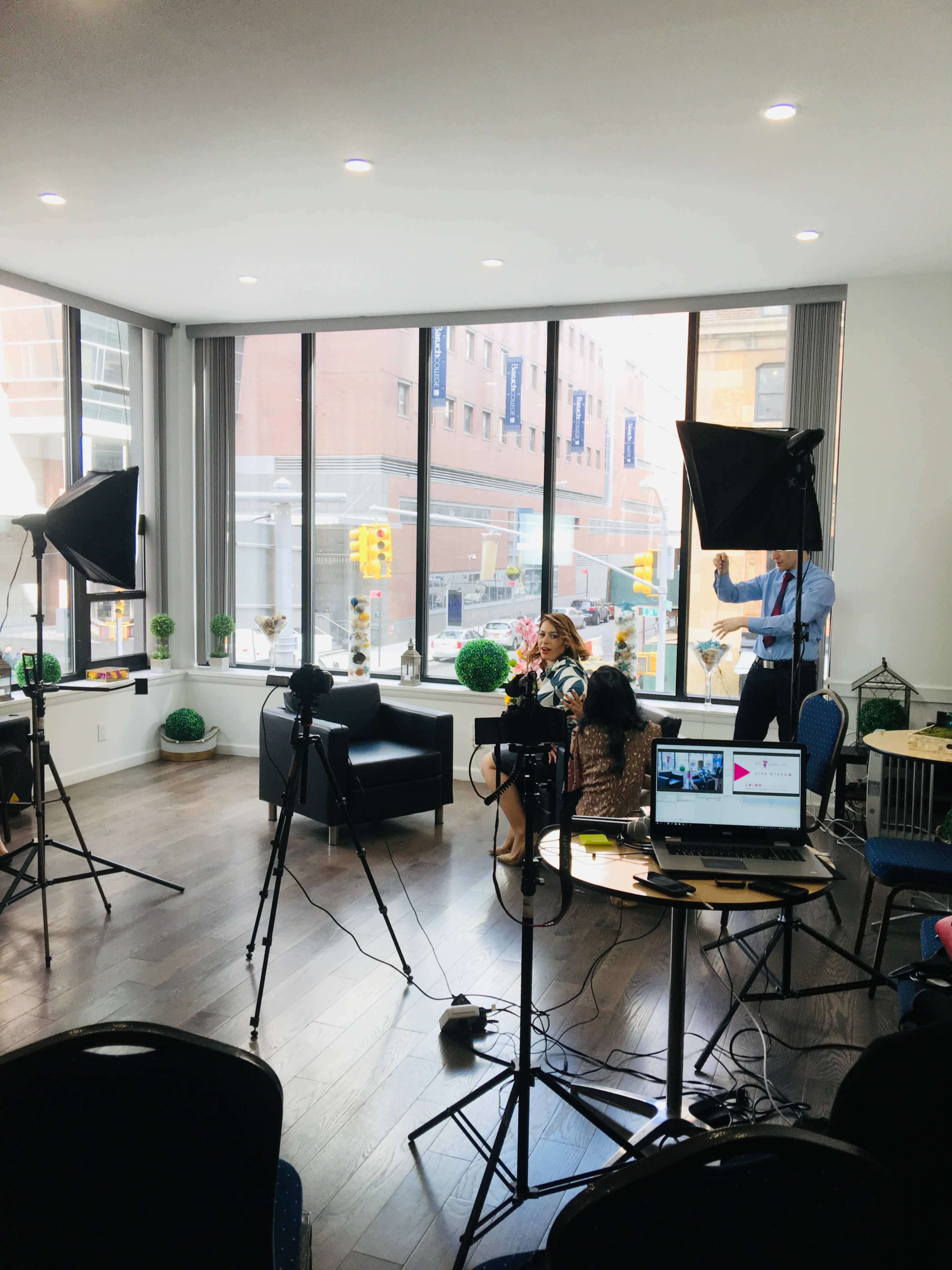 A well-lit interview setup with cameras, lighting equipment, and a laptop on a table, overlooking a city street through large windows.