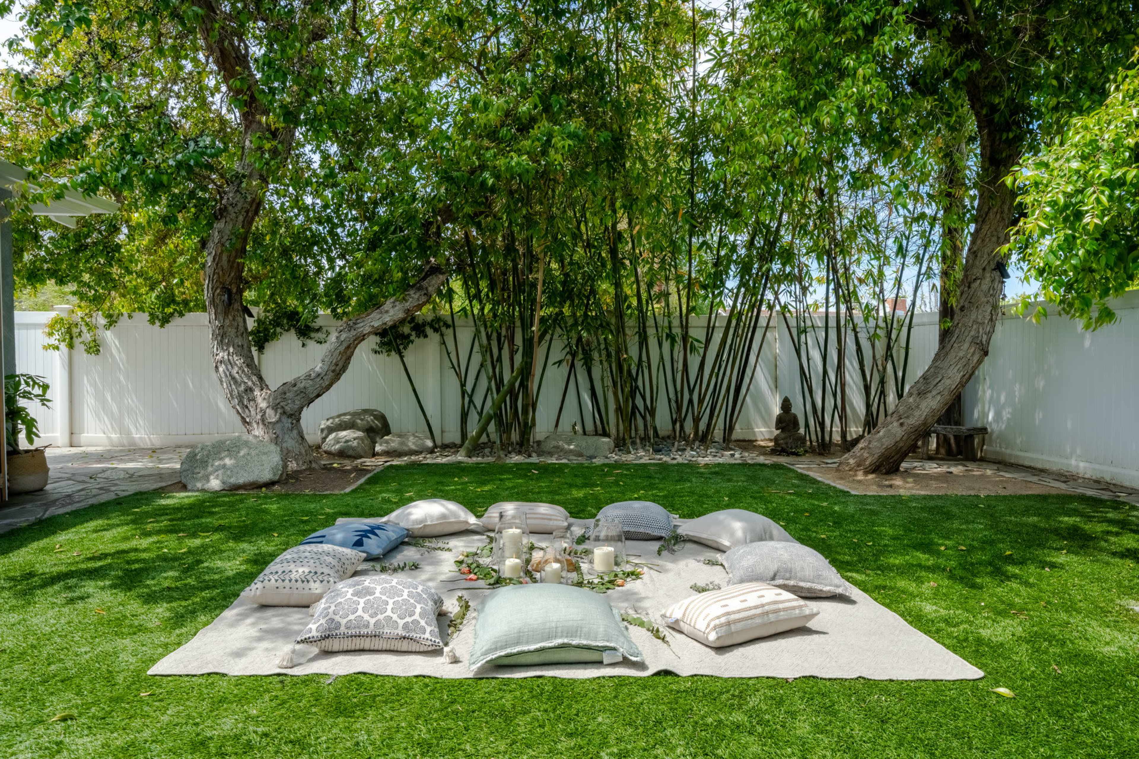 A picnic setup with various cushions and decorative items is arranged on a grassy area surrounded by trees and a white fence.
