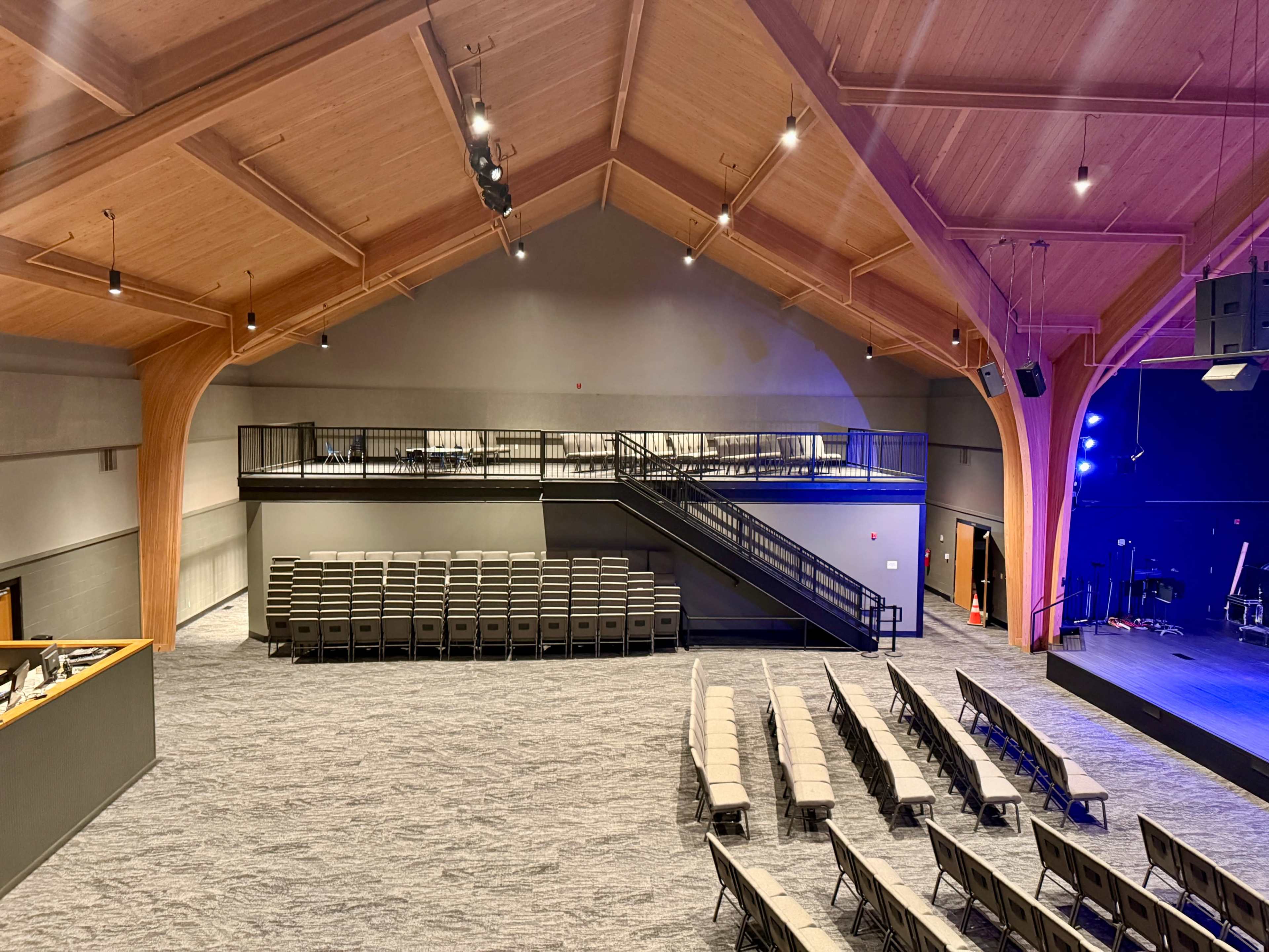 The image shows the interior of a spacious auditorium with tiered seating, wooden beams on the ceiling, and a stage area equipped with lighting.