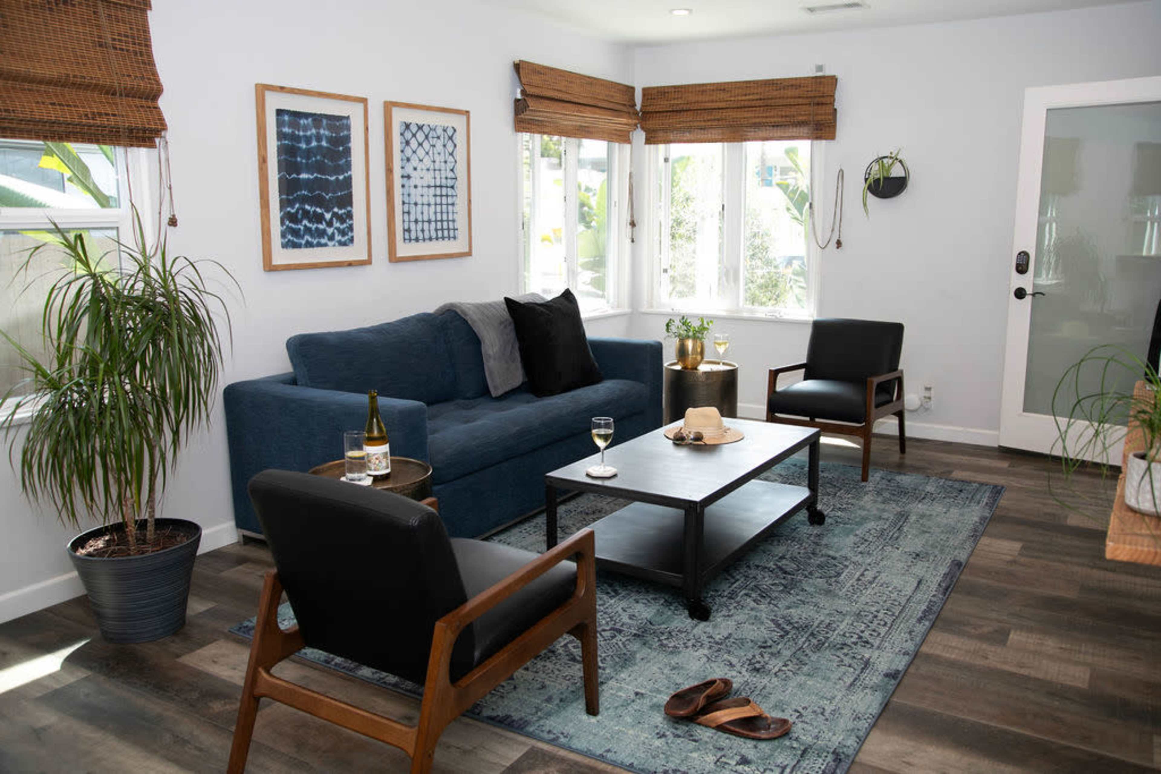The living room features a blue sofa, a coffee table, two black chairs, and a potted plant, all set against light-colored walls and large windows with bamboo shades.