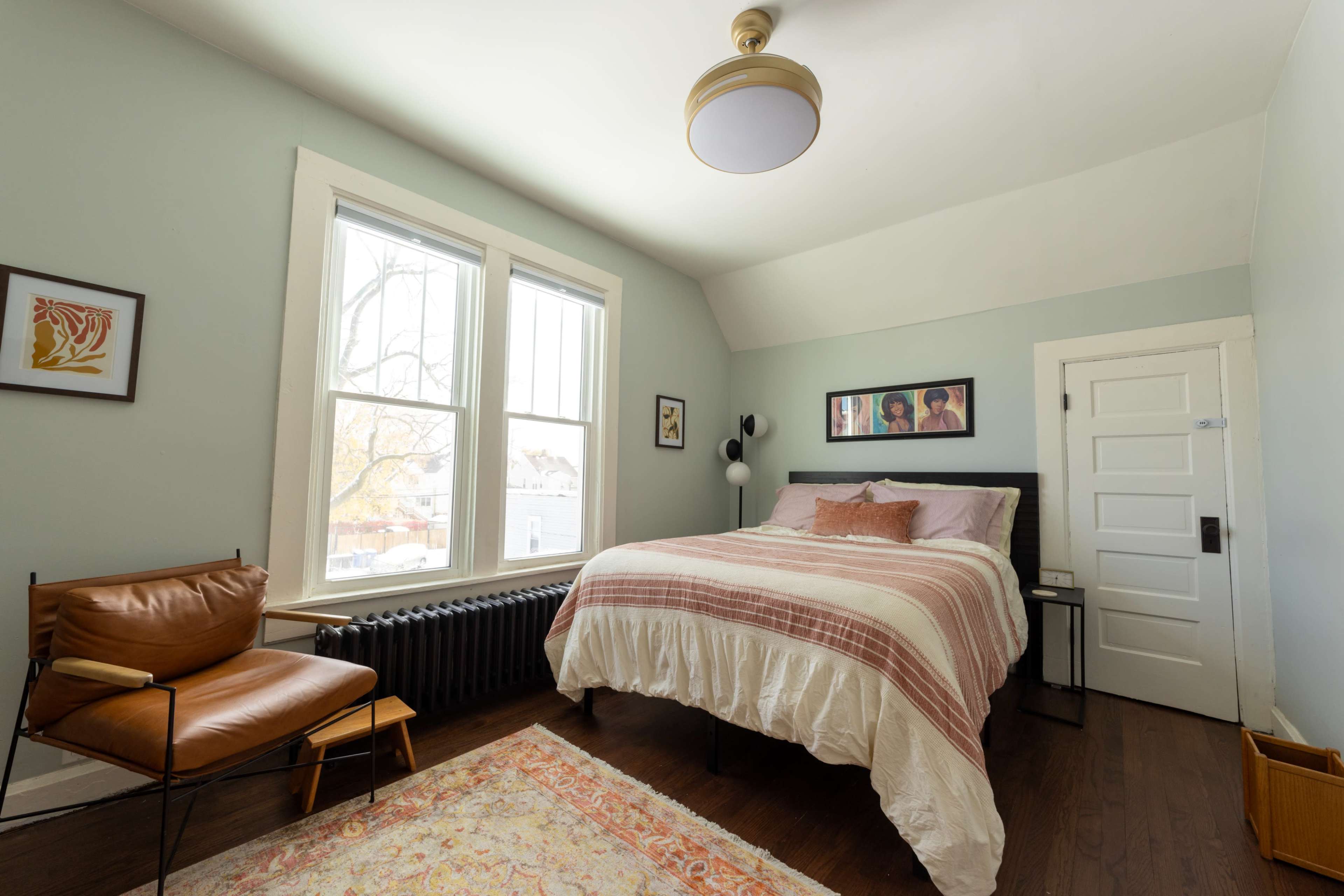 The image shows a cozy bedroom with a large bed, a vintage chair, and windows letting in natural light.
