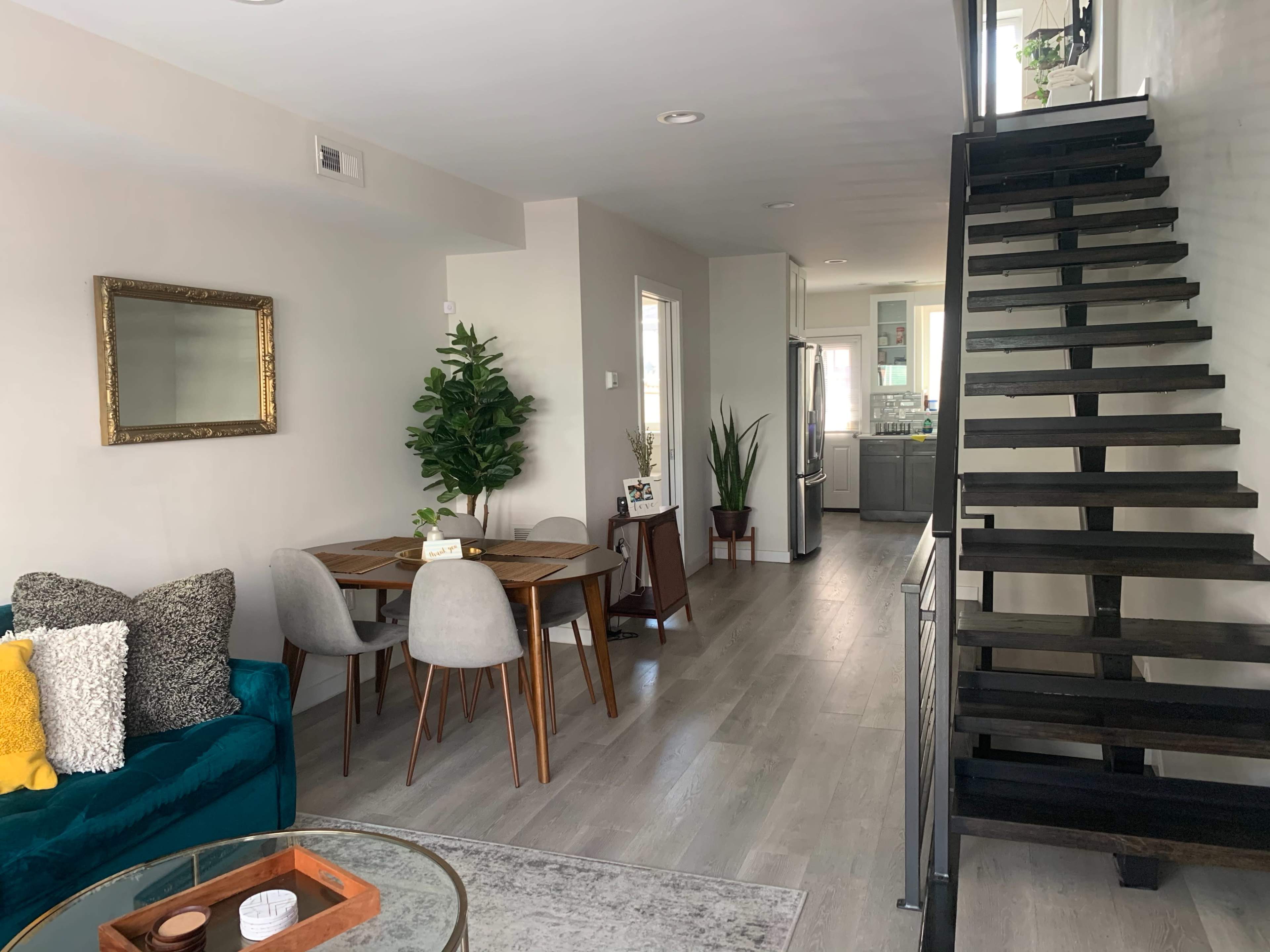 The image shows a modern, open-plan living area featuring a dining table, plush seating, a staircase, and kitchen area in the background.