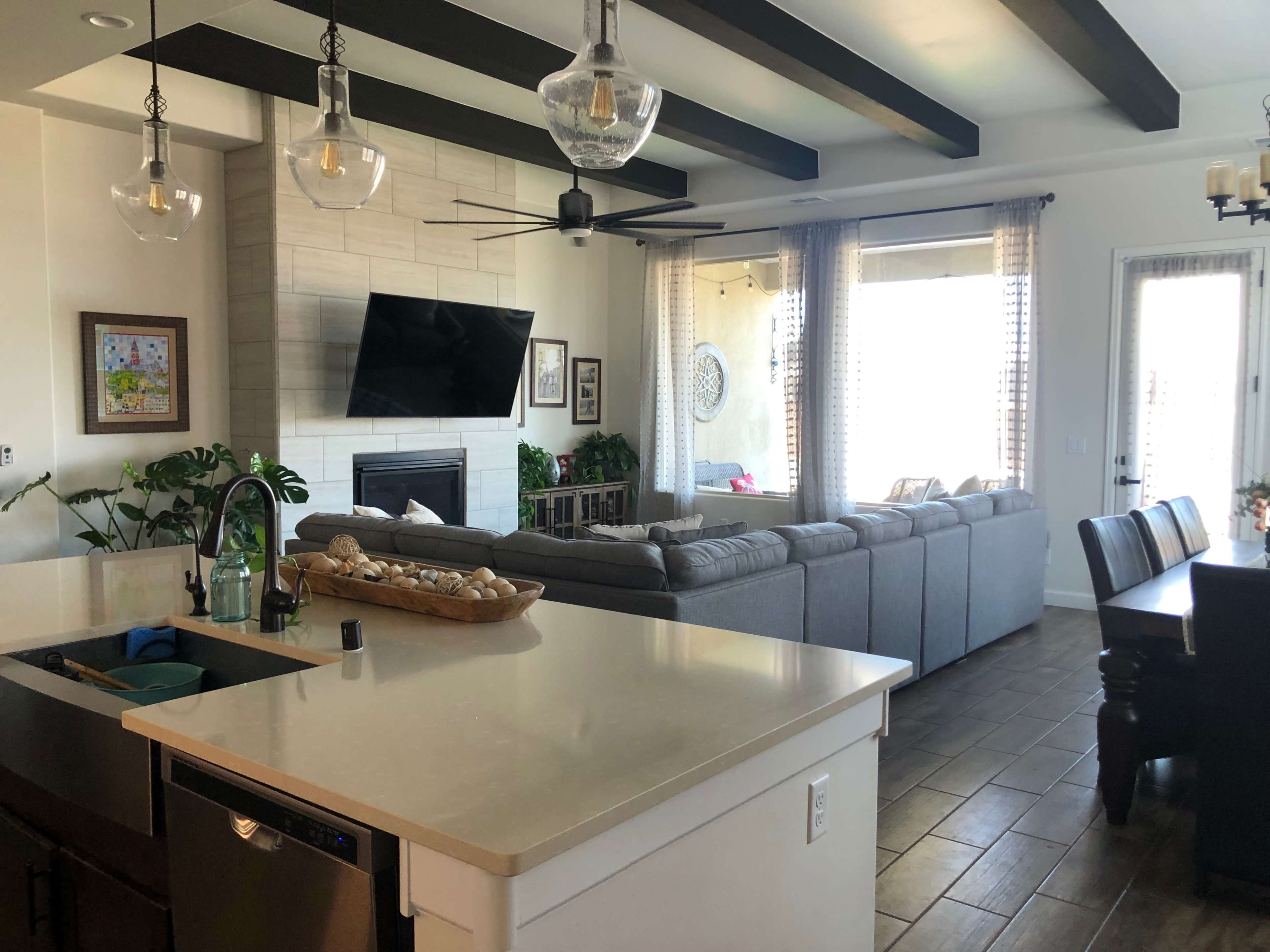 The image shows a modern living space featuring a kitchen area with a countertop, a living room with a large gray sofa, and a dining area with a wooden table, all illuminated by natural light.