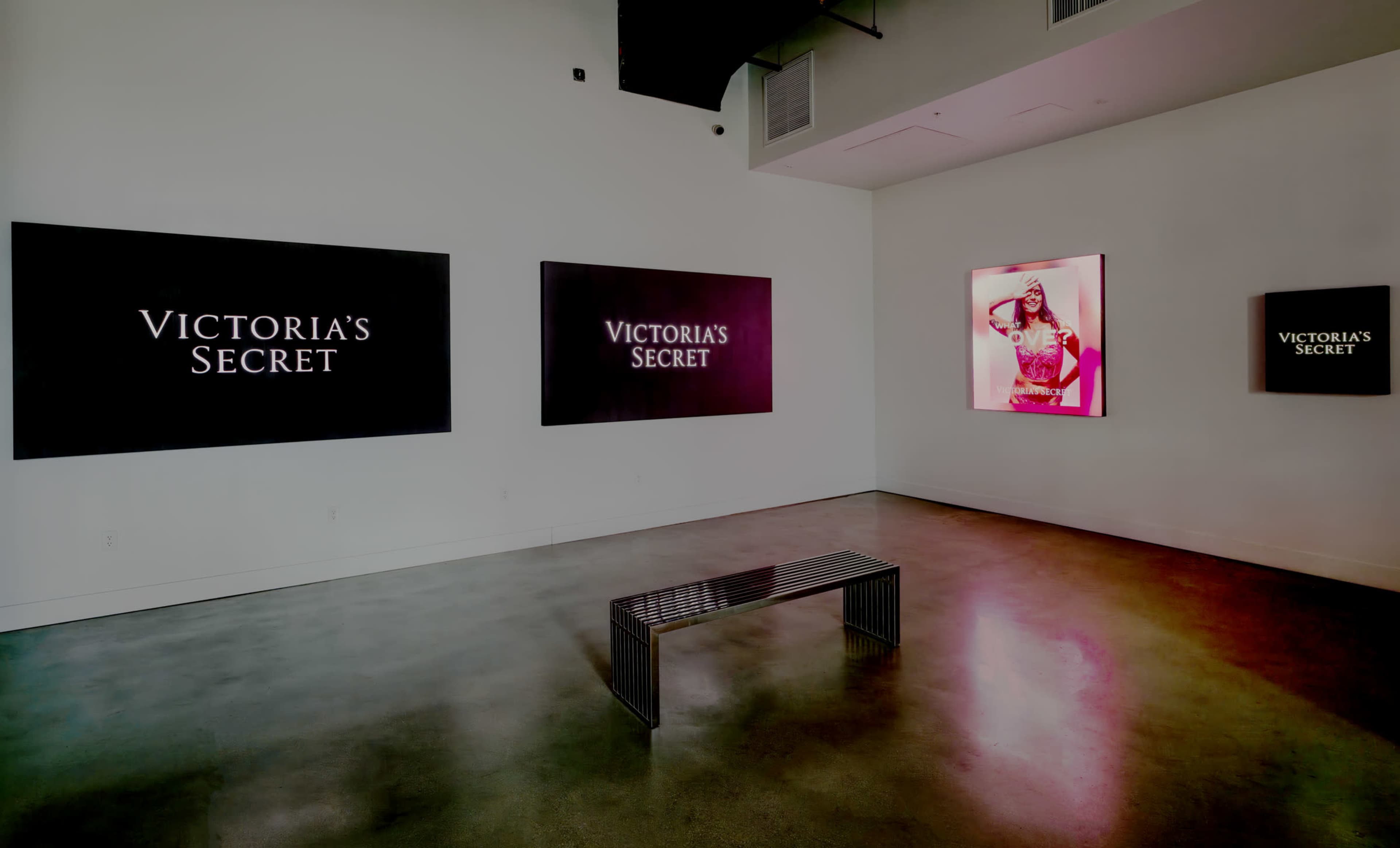 The image shows an art gallery with dark walls displaying "Victoria's Secret" in large text on multiple screens, alongside a framed photograph of a model.