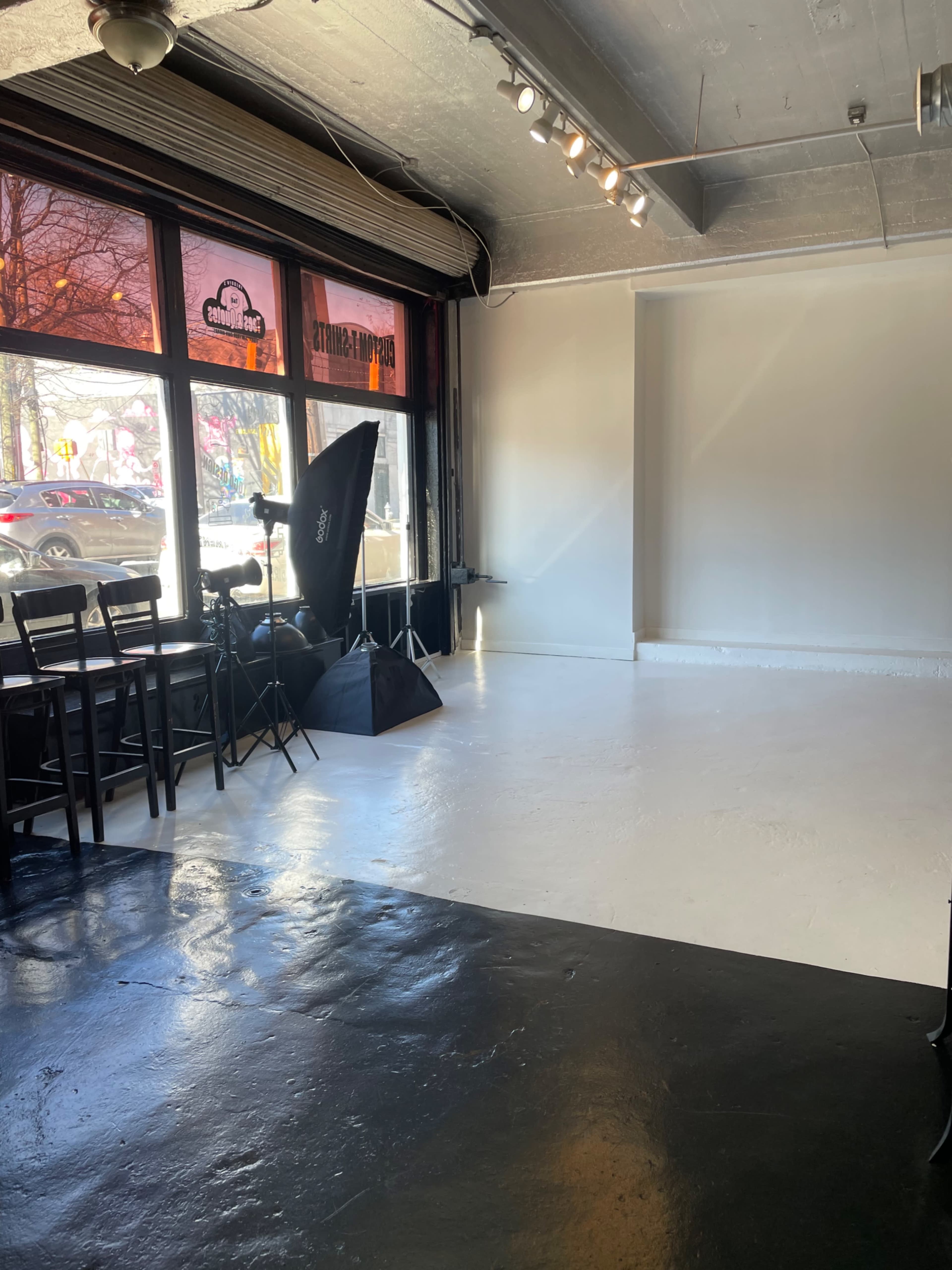 A spacious interior with a white floor, black chairs, and a lighting setup in front of large windows.