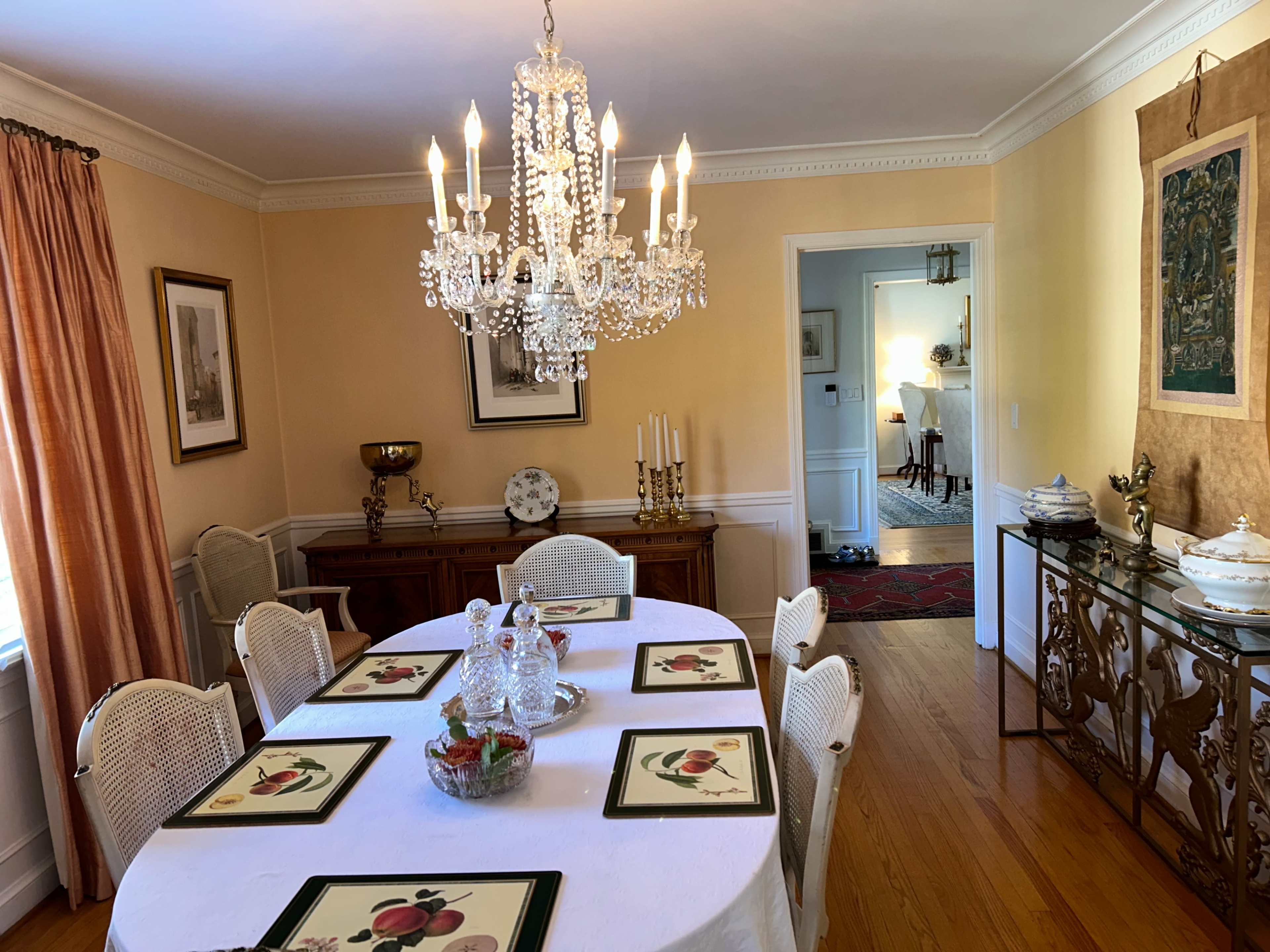 The dining room features a round table set with placemats and surrounded by white chairs, illuminated by a chandelier, with a sideboard and decorative pieces against the walls.
