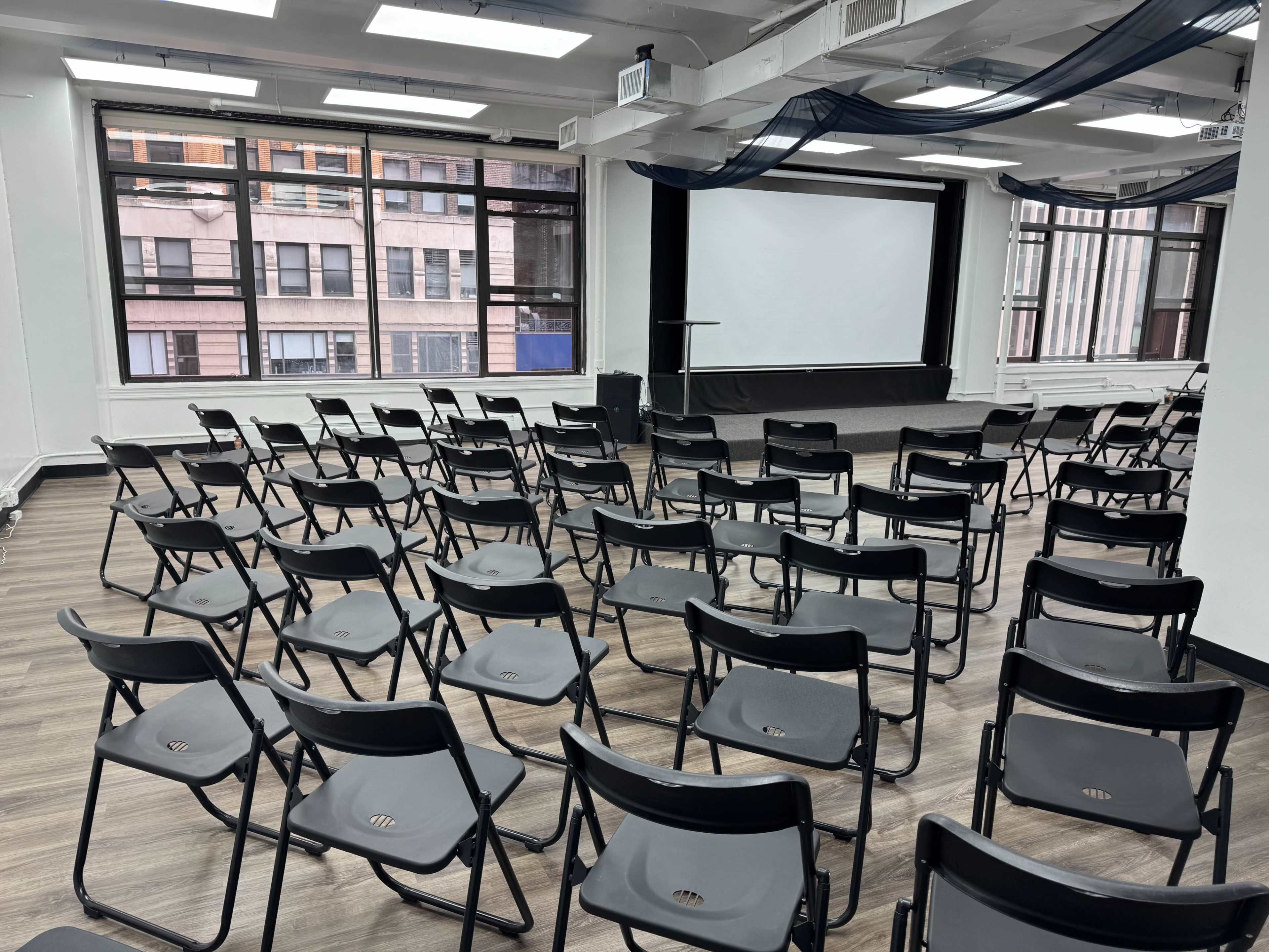 A large room is set up with rows of black folding chairs facing a white screen at the front, with large windows providing natural light.