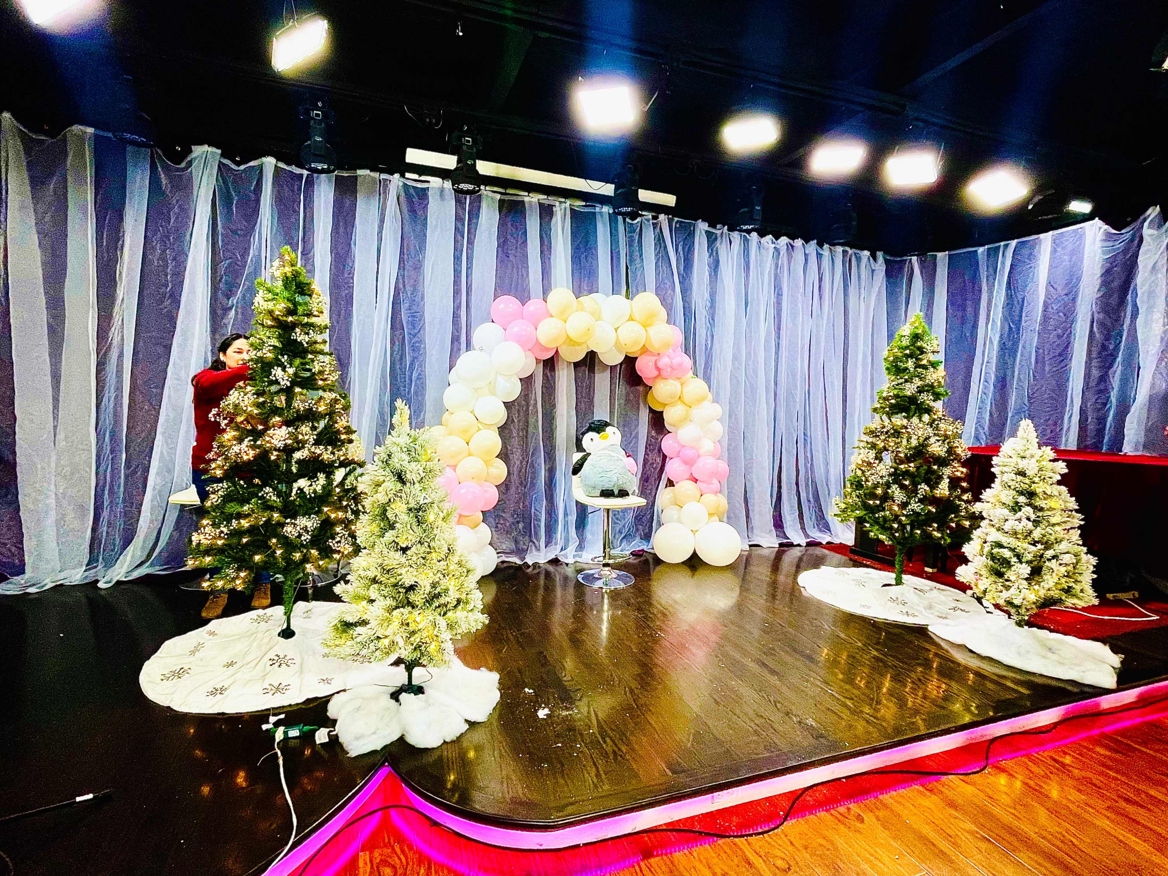 The scene features a stage decorated with three Christmas trees, an arch of balloons, and a plush figure seated on a stool.