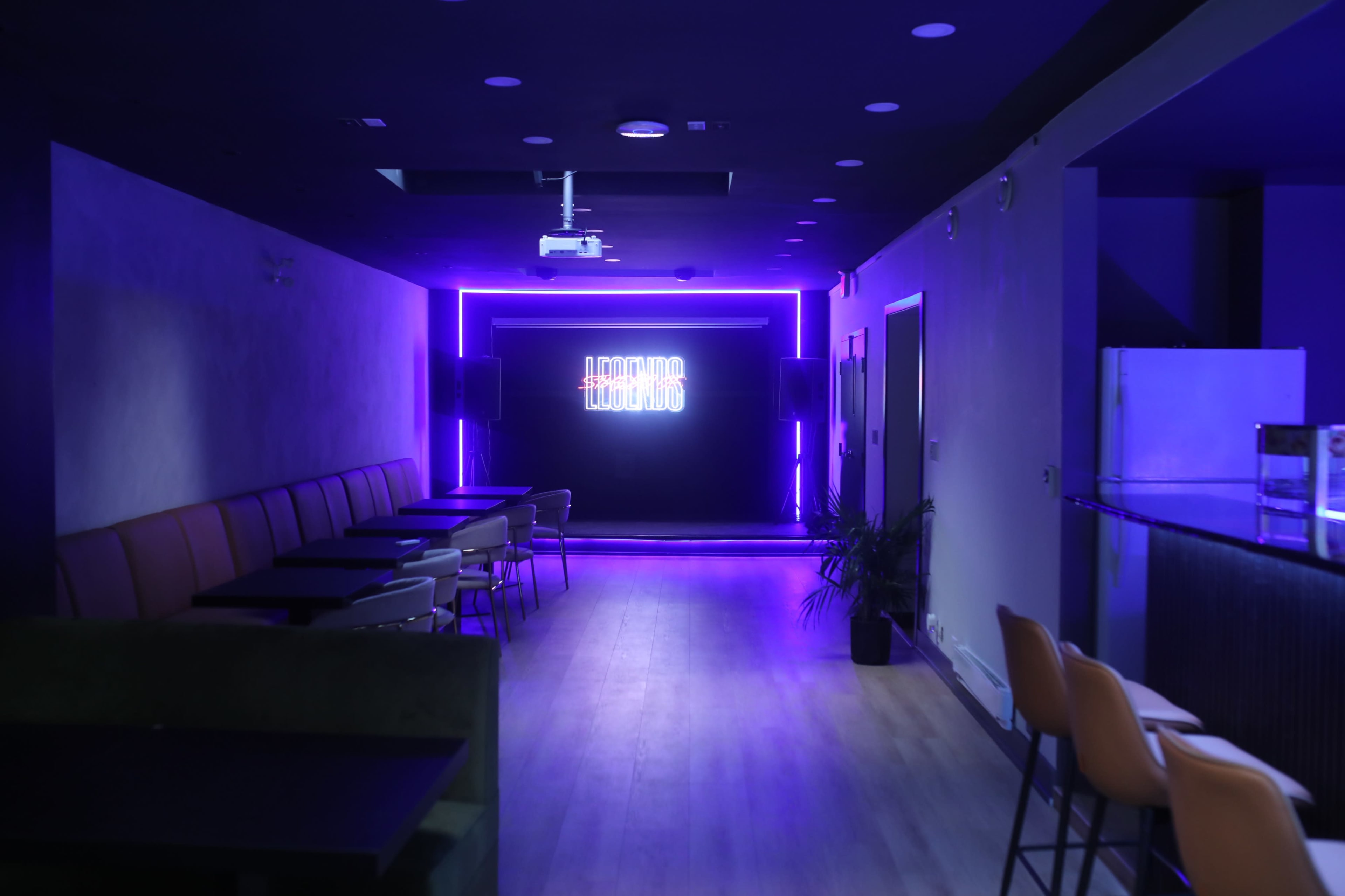 The image shows a modern lounge with purple neon lighting, featuring rows of tables and chairs along the walls.