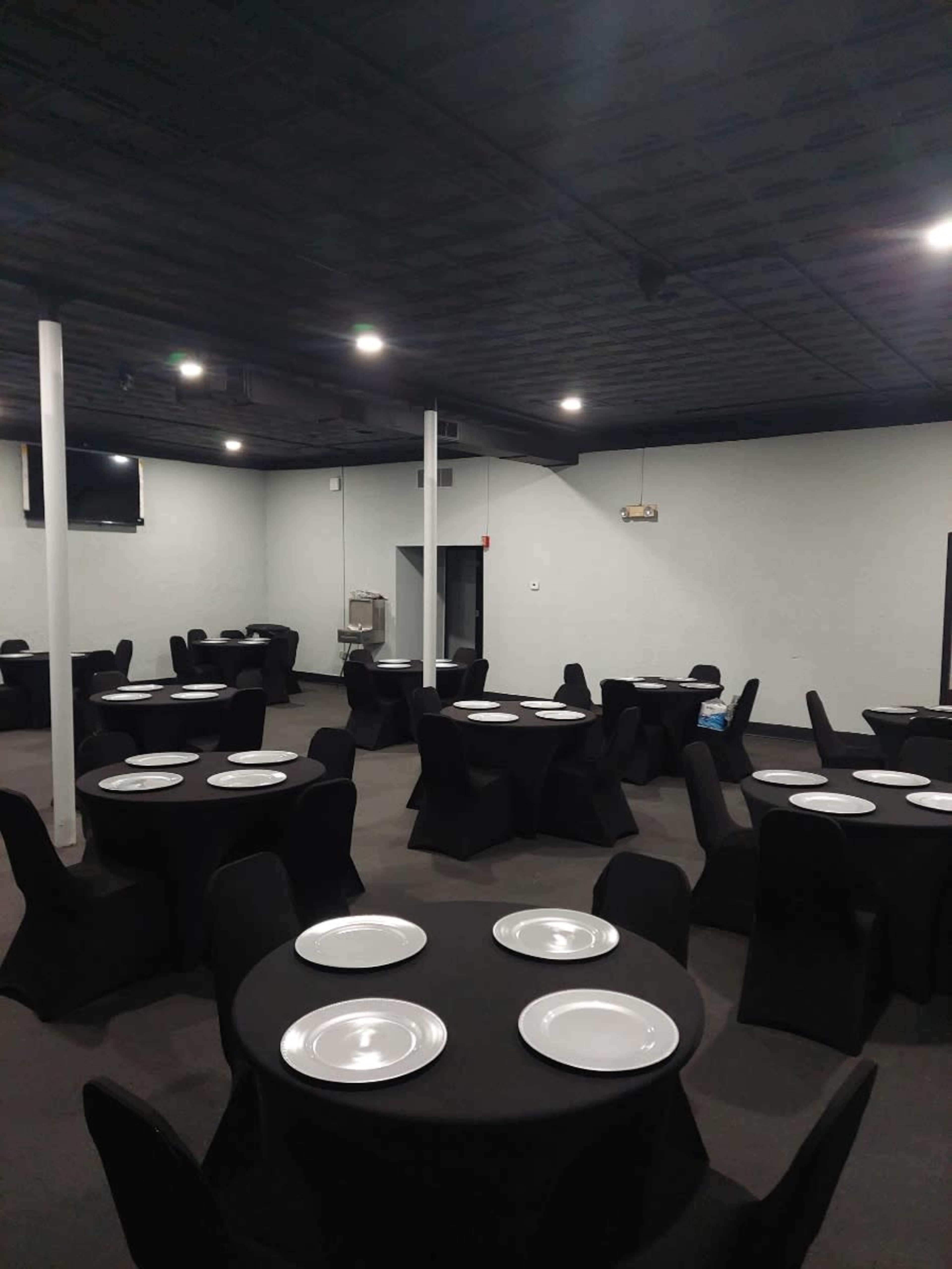 The image shows a spacious dining event room with round black tables, each set with white plates, illuminated by overhead lights.