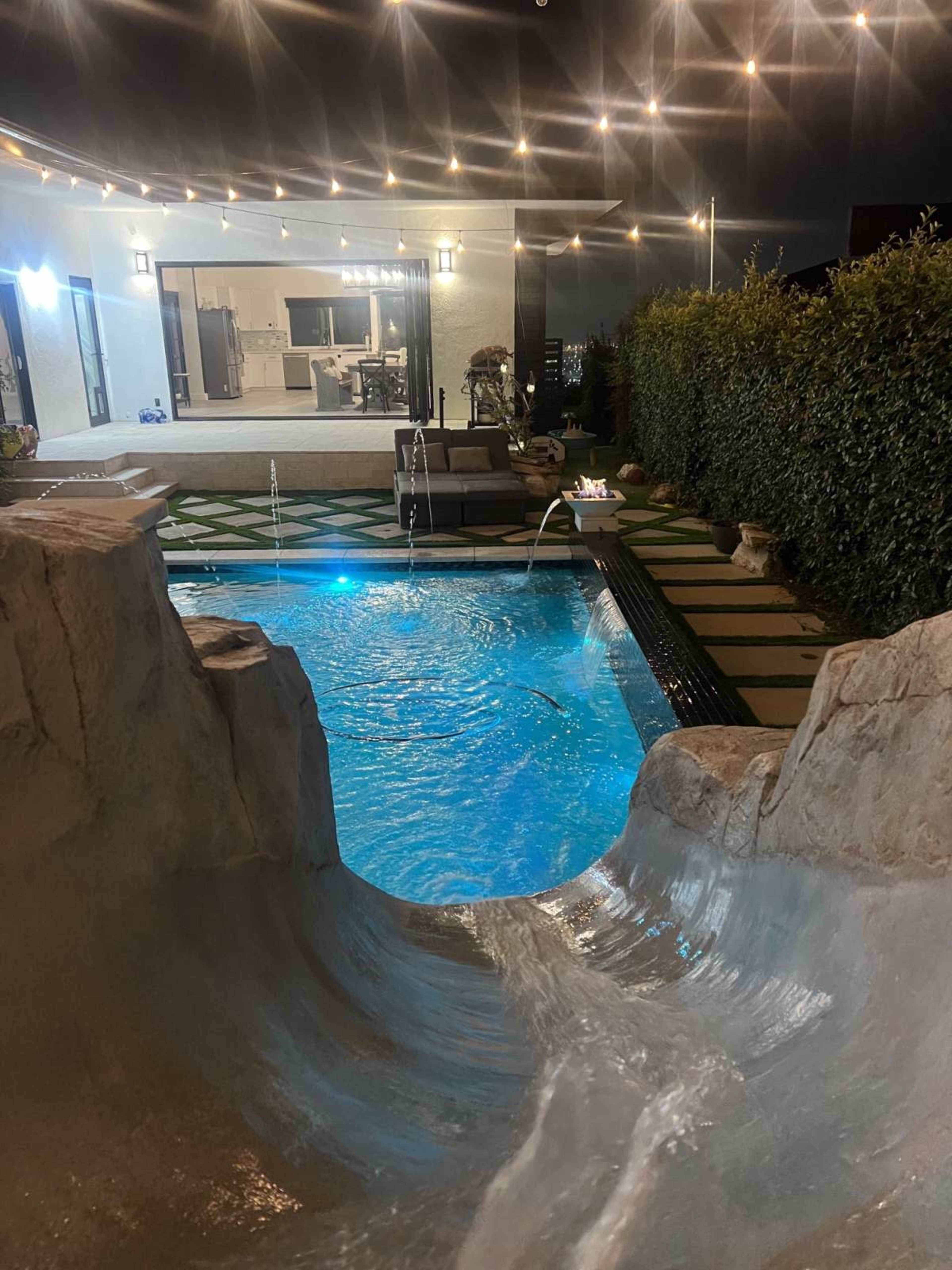 The image shows a swimming pool with a water slide and a modern home illuminated by outdoor string lights in the background.